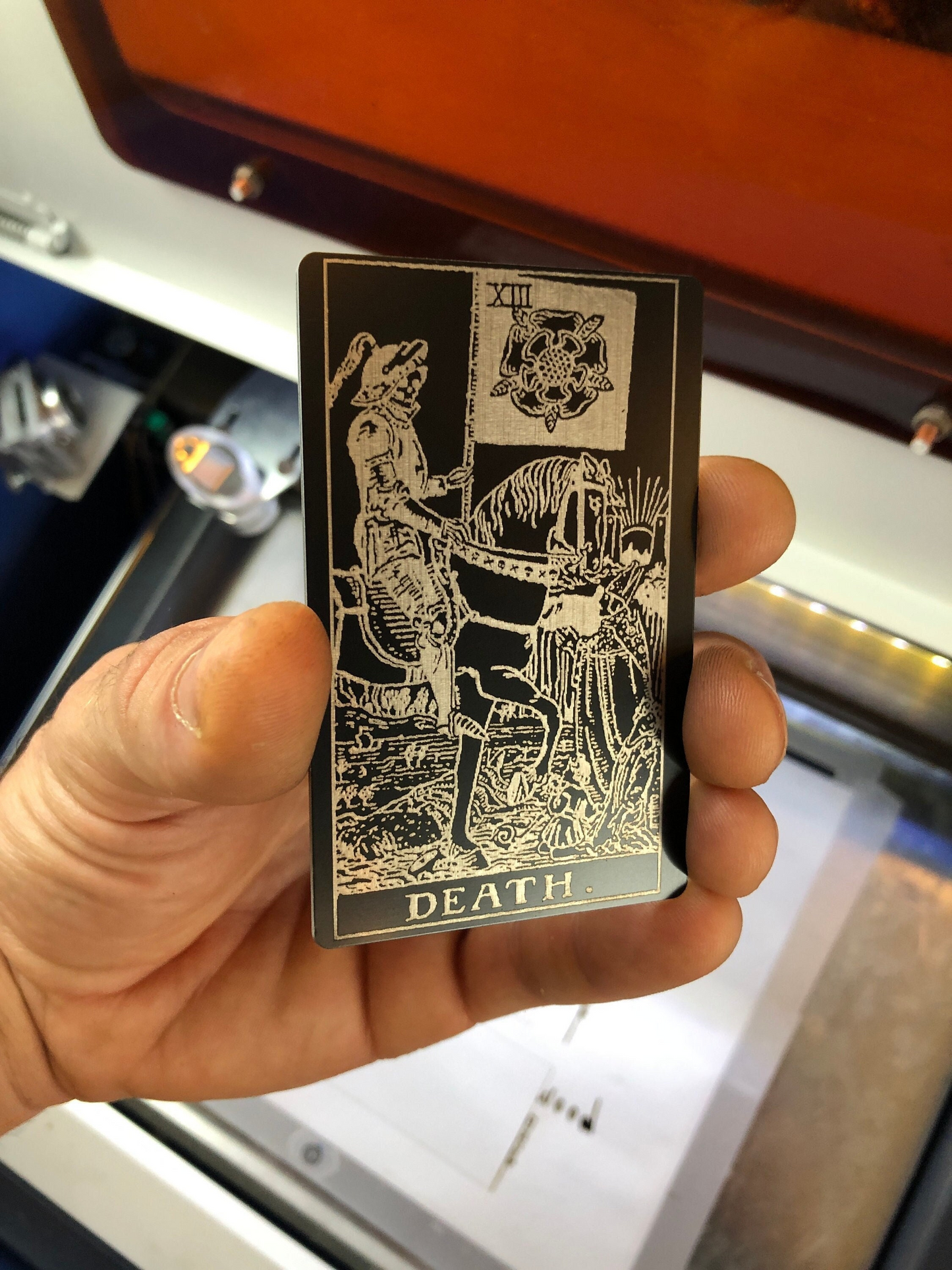 Hand holding a metal tarot card keepsake with 'Death' symbol against a blurred indoor background