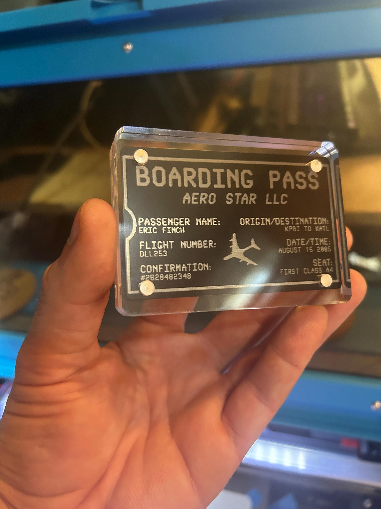 Hand holding a transparent acrylic display case with a metal laser engraved keepsake inside reading 'Boarding Pass' text and branding