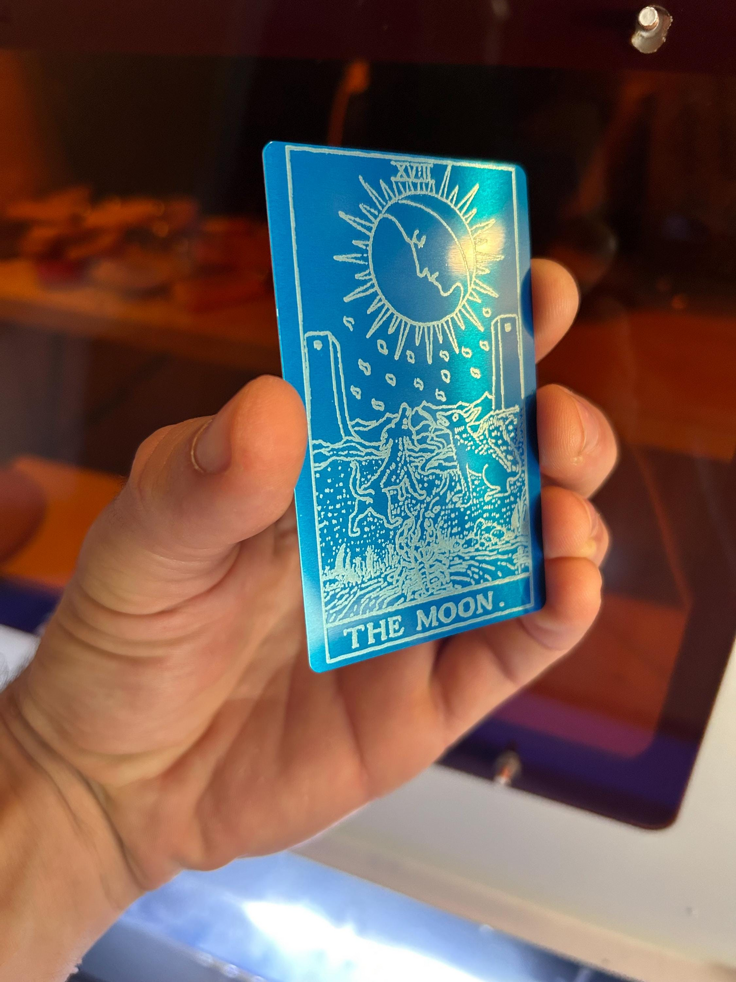 Hand holding a blue and silver metal tarot card titled 'The Moon'