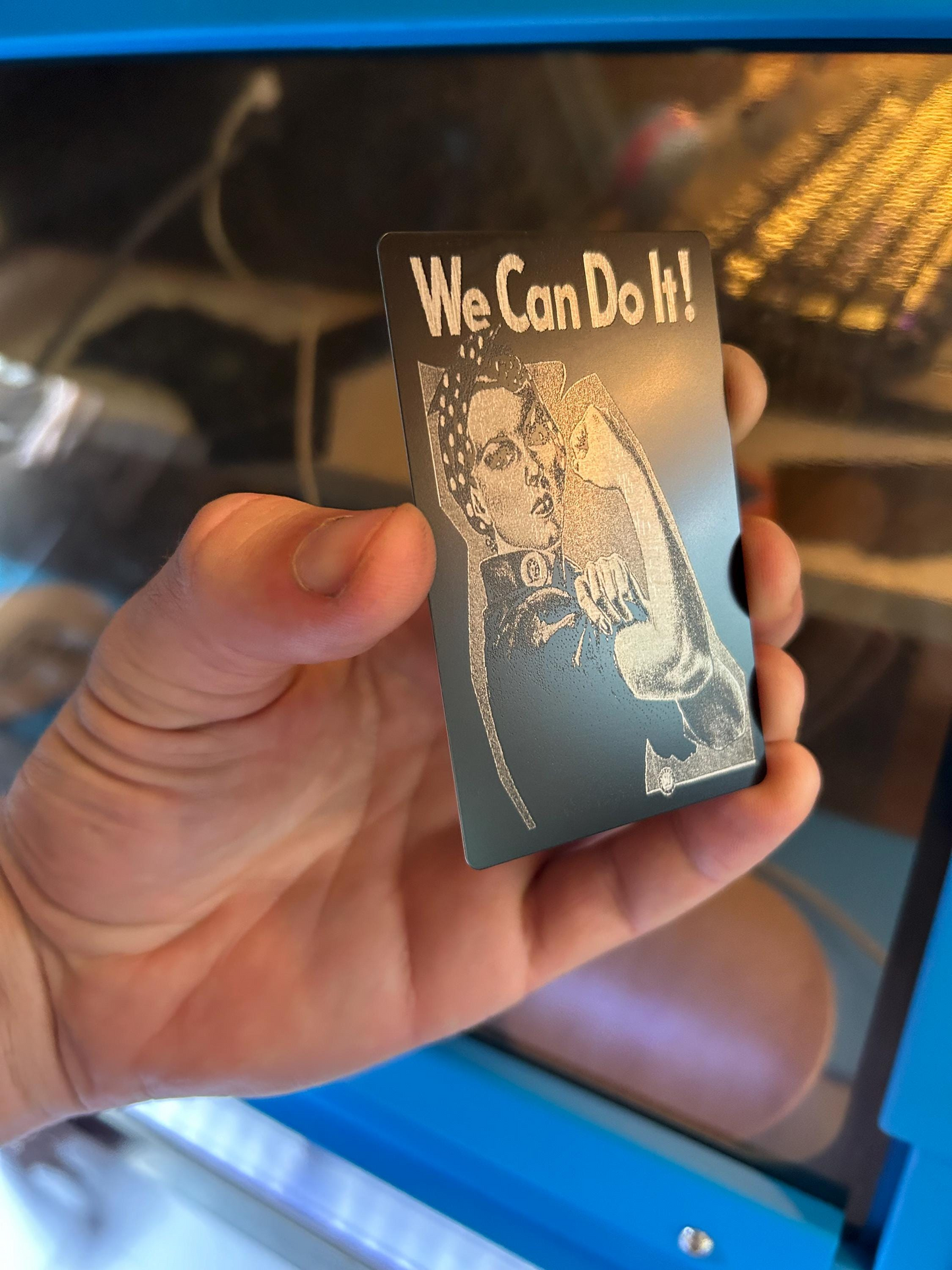 Hand holding a metal laser engraved card with 'We Can Do It!' text and a vintage-style illustration.