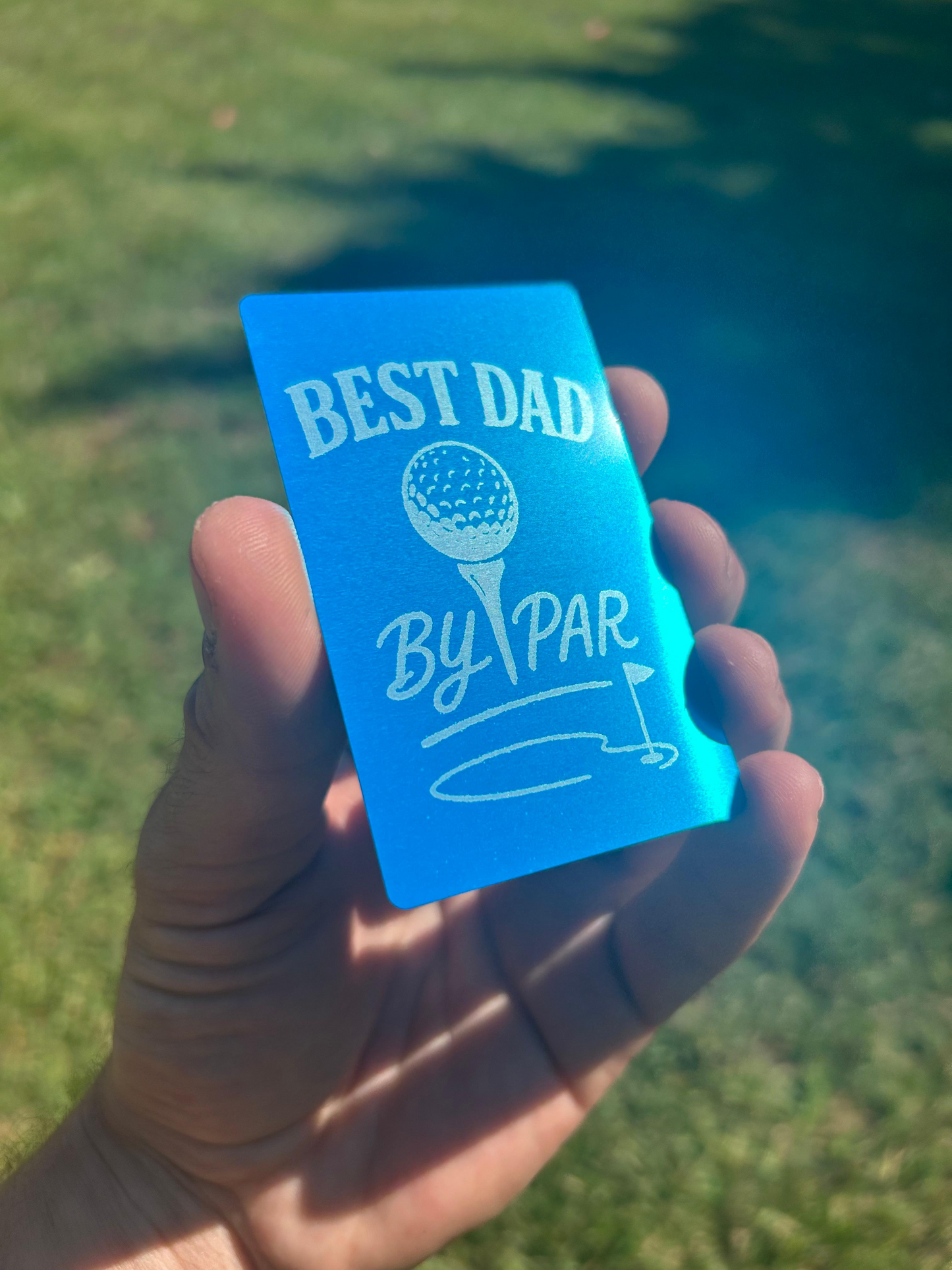 Hand holding a blue/silver metal engraved keepsake card with 'Best Dad by Par' text and golf graphics against a blurred green background.