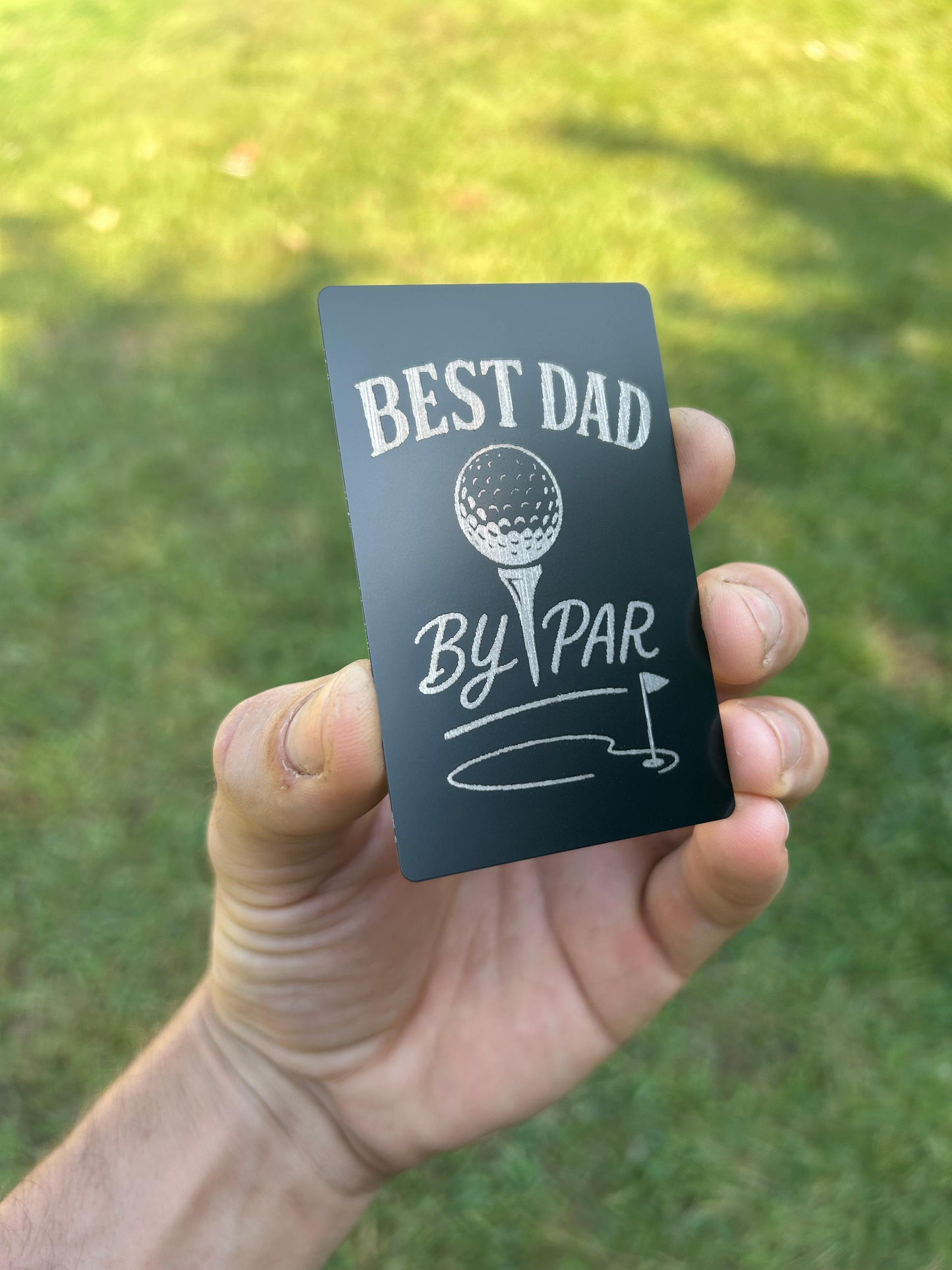 Hand holding a metal laser engraved card with 'Best Dad by Par' text against a grassy background