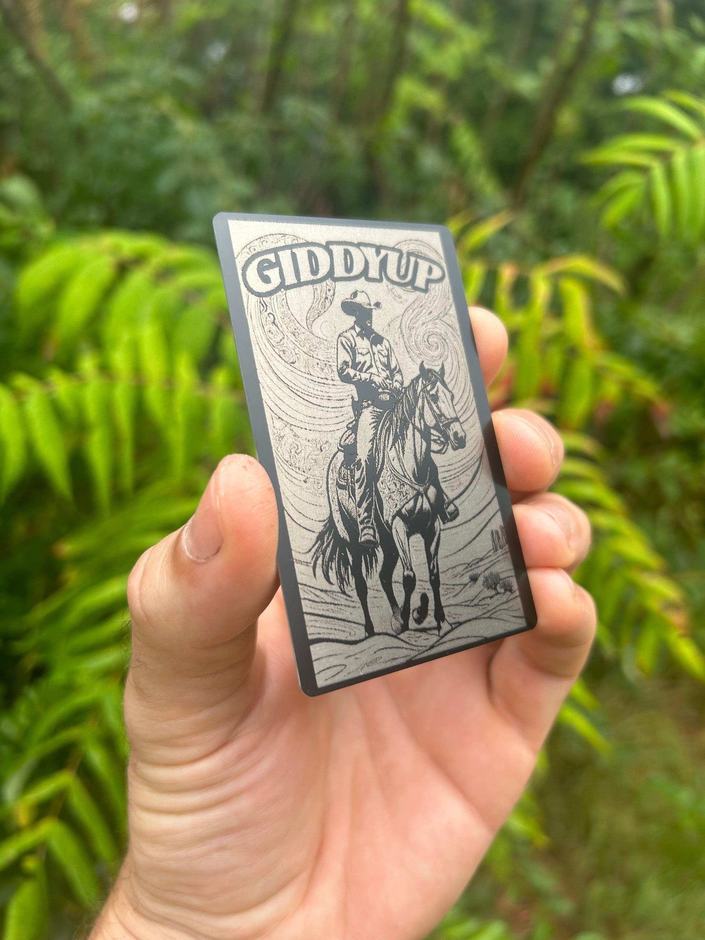 Hand holding a metal Cowboy 'Giddy Up' card with a horse and rider illustration against a green background