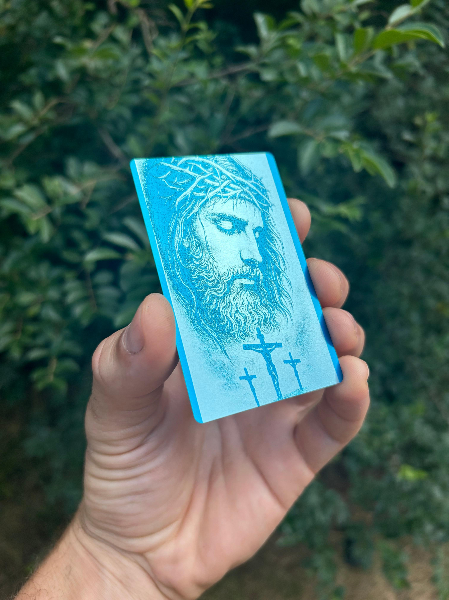 Hand holding a blue/silver laser engraved metal bookmark card with a christian inspirational design against a green leafy background
