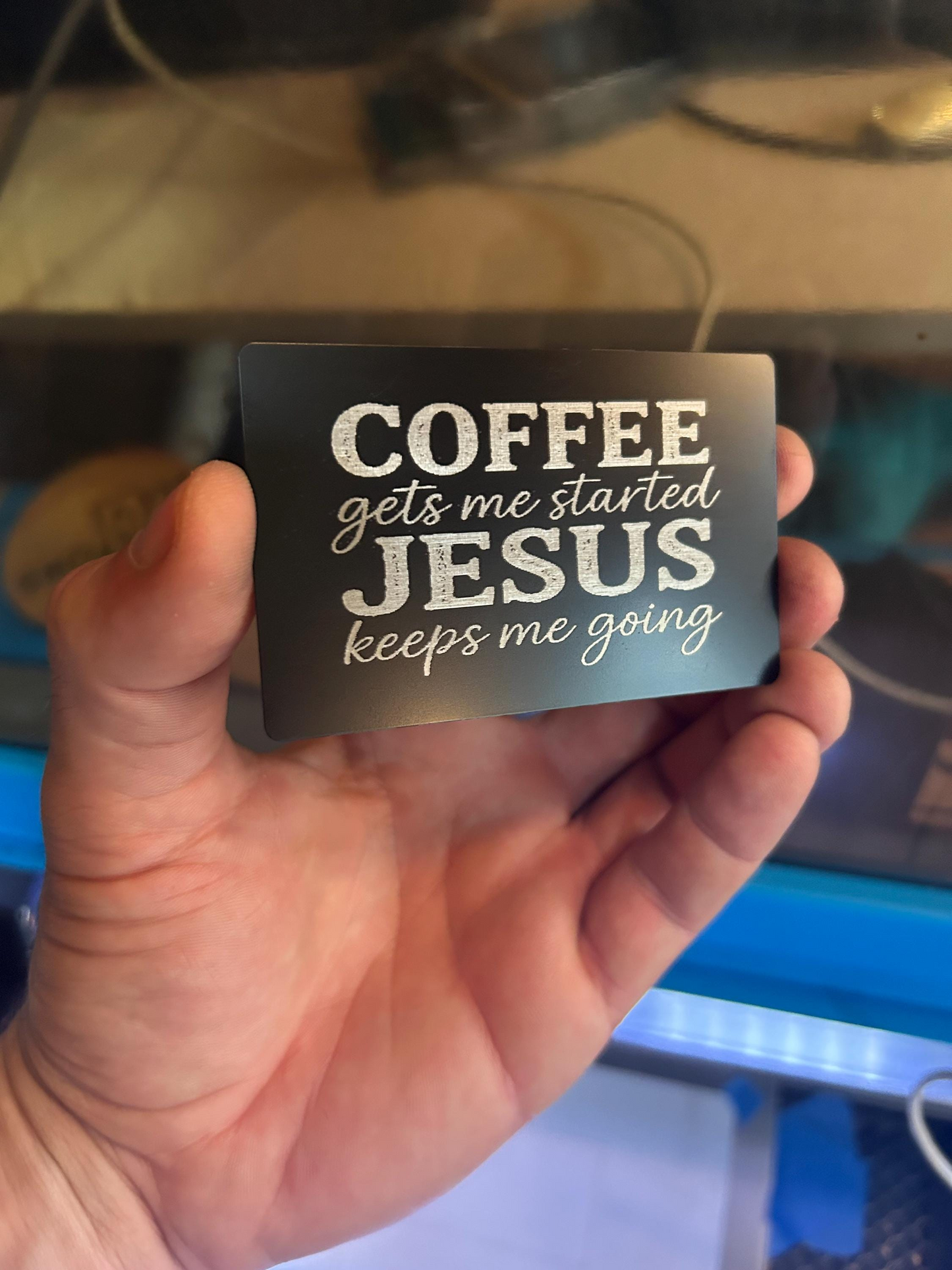 Hand holding a metal christian gift card with text 'COFFEE gets me started JESUS keeps me going' in black/silver