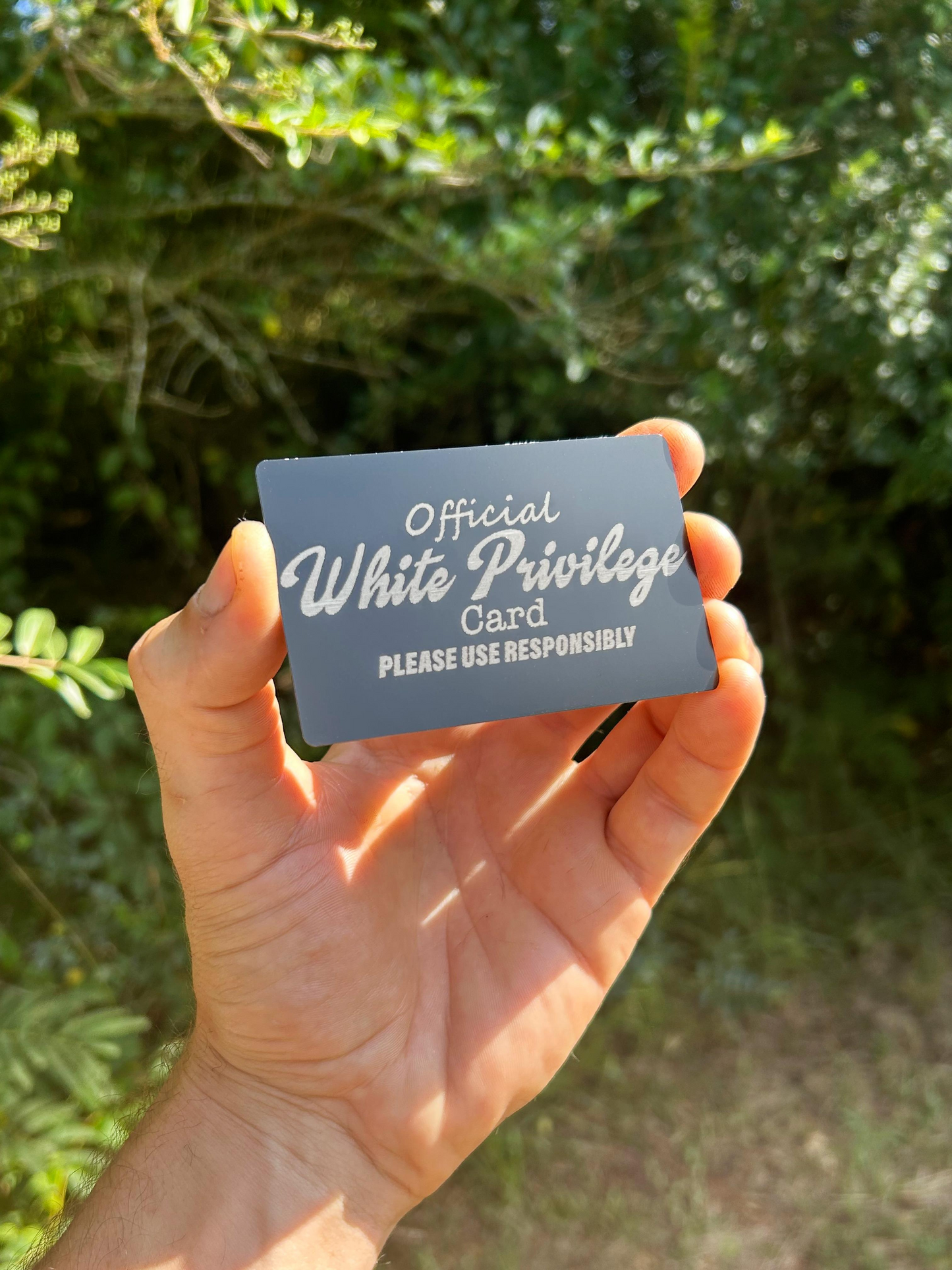 Hand holding a metal keepsake gag gift card with 'Official White Privilege Card' text laser engraved against a blurred green background