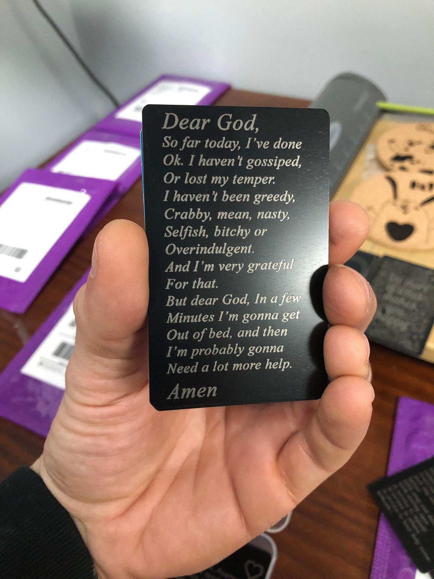 Hand holding a black metal prayer card with custom engraved text on it, with a blurred shipping department background