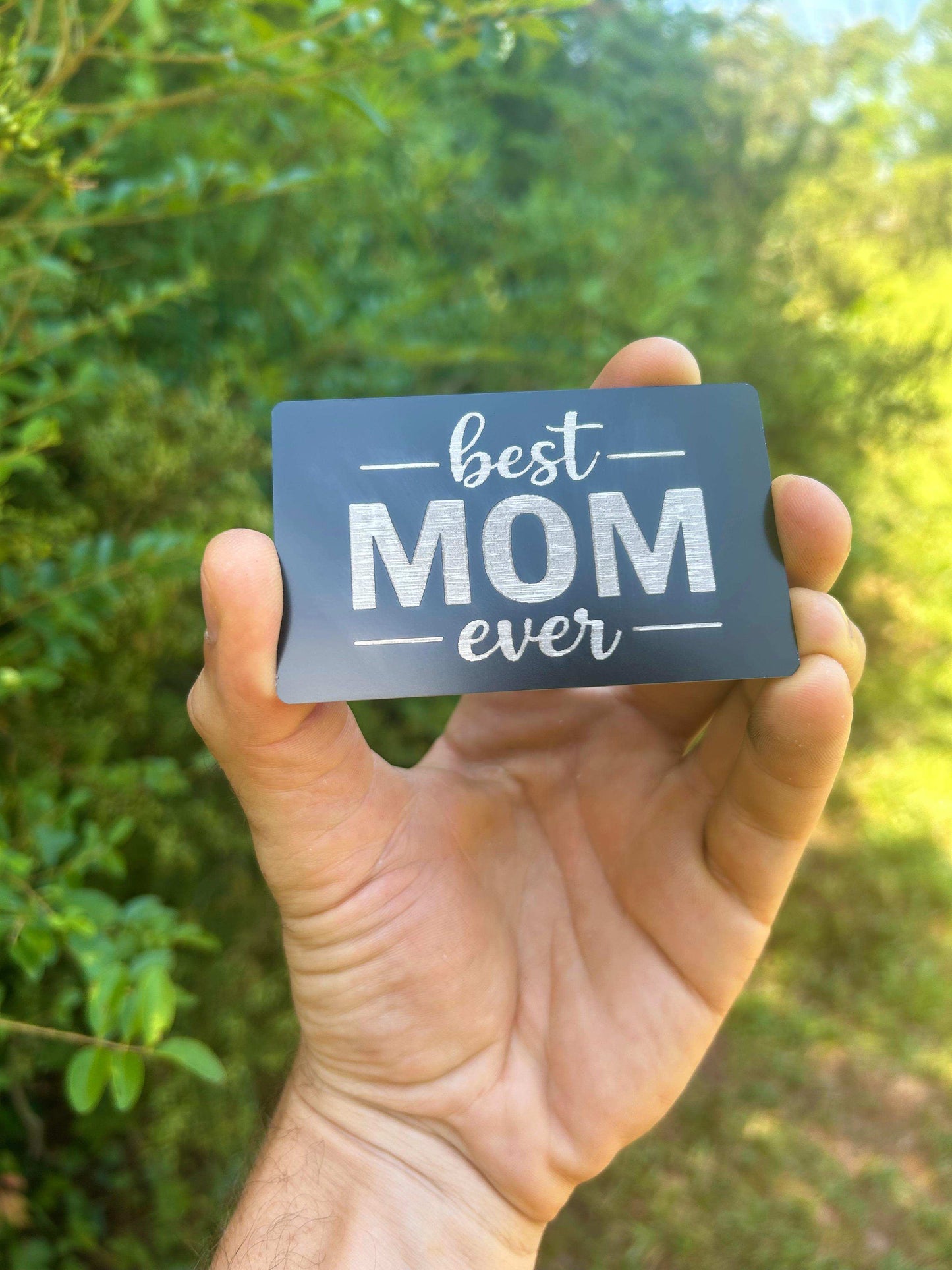 Hand holding a metal mother's day card with 'best mom ever' text against a blurred green outdoor background