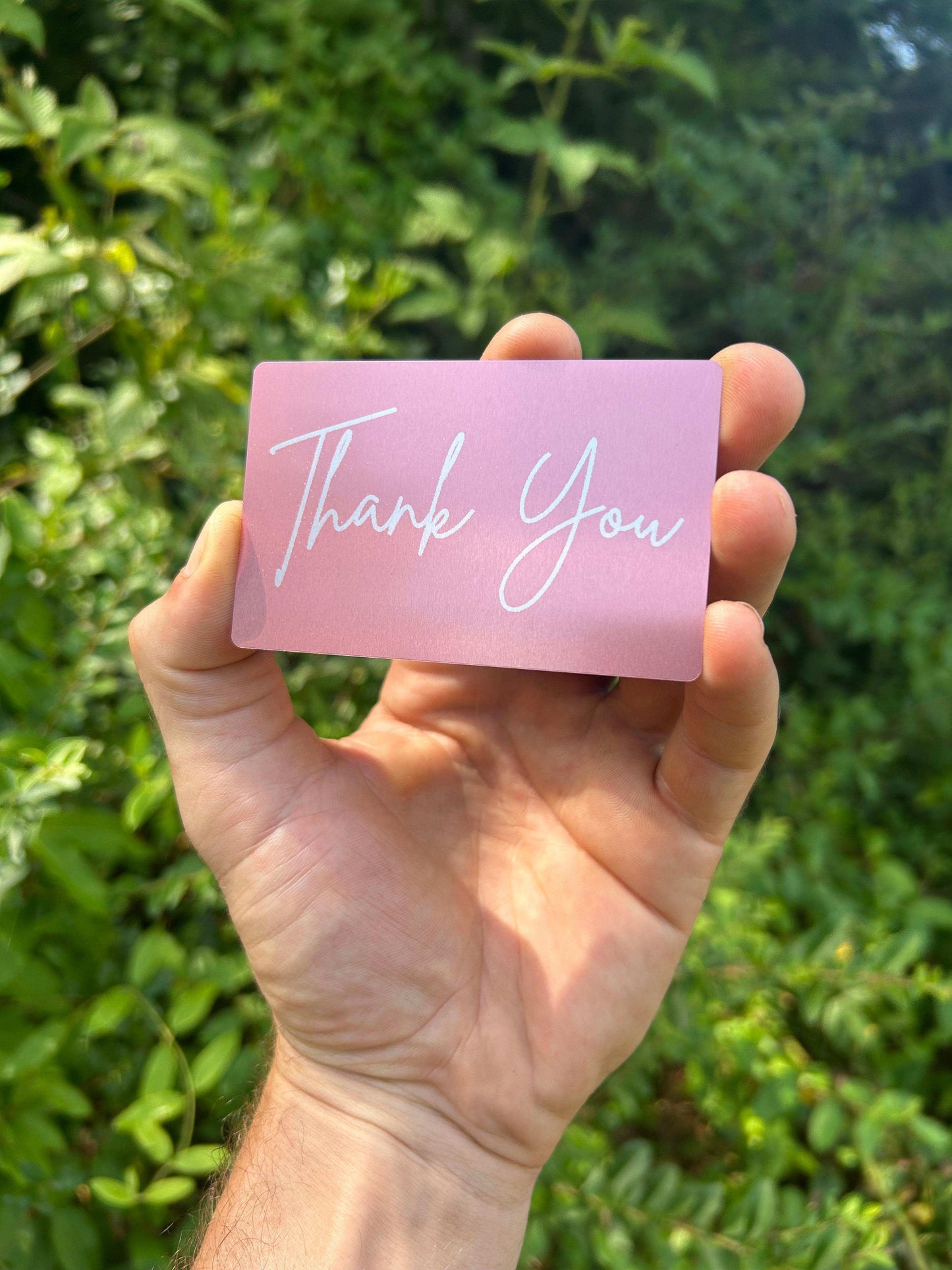 Hand holding a pink card with 'Thank You' text against a green leafy background