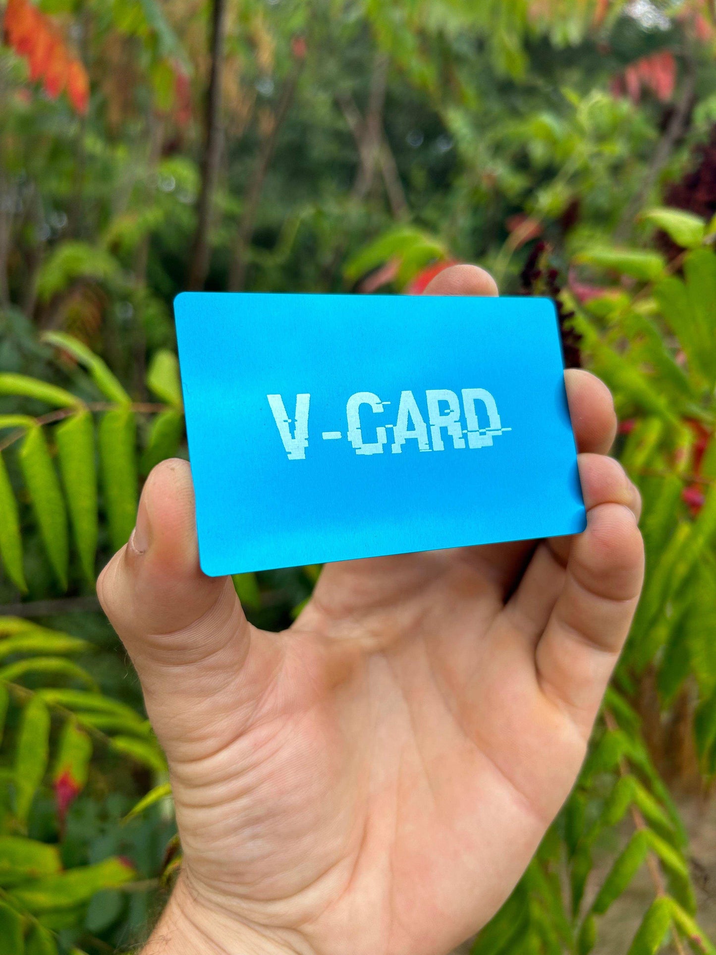Hand holding a blue metal card engraving with 'V-CARD' text against a green background