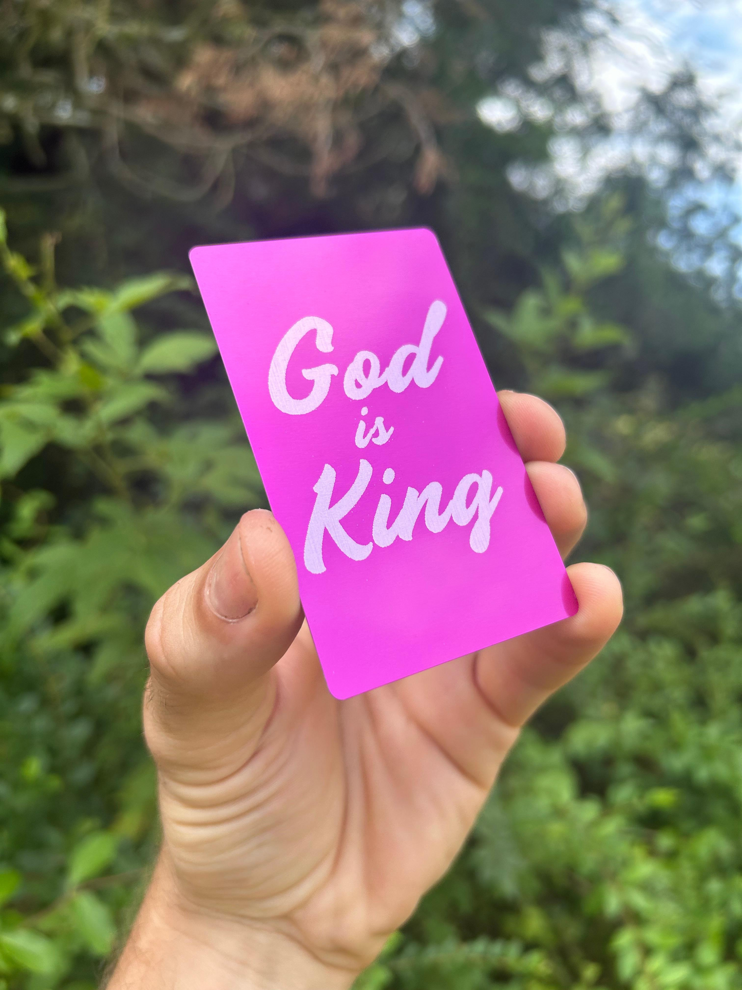 Hand holding a purple card with 'God is King' text against a blurred green outdoor background
