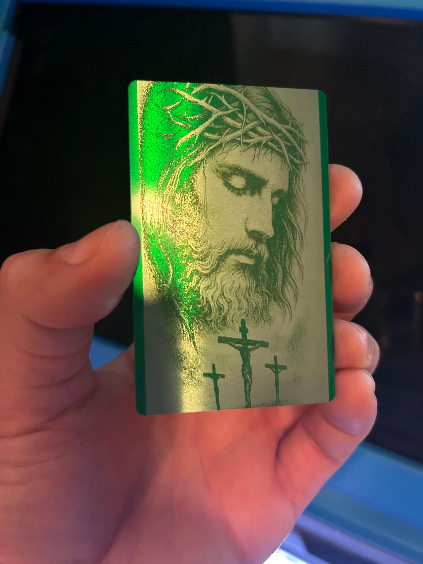 Hand holding a metal green card with a religious design featuring jesus and crucifix.