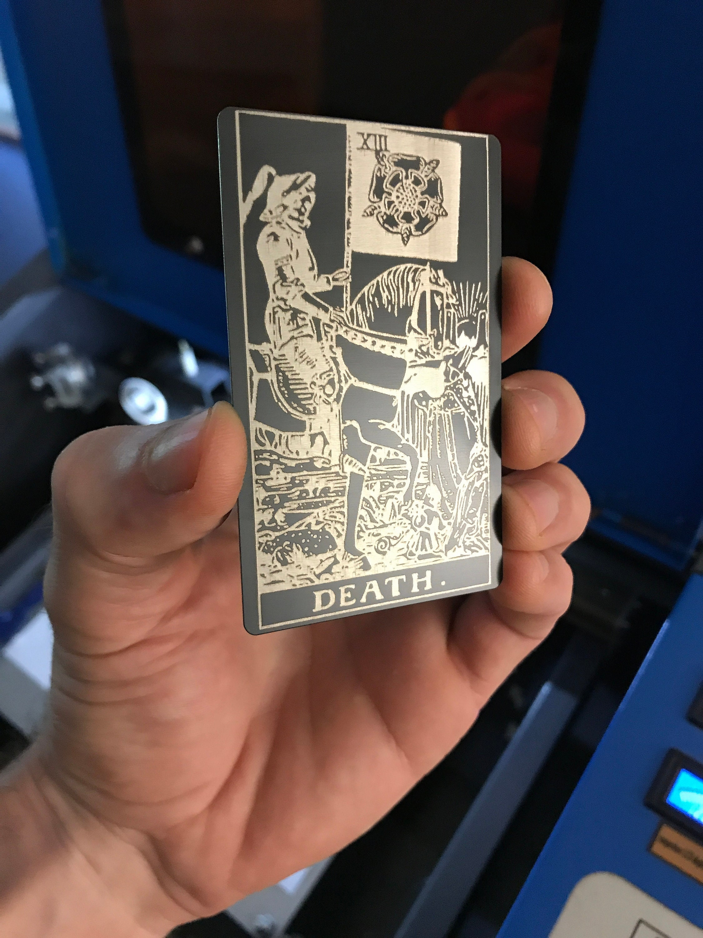 Hand holding a matte black/silver metal tarot card with 'Death' symbol