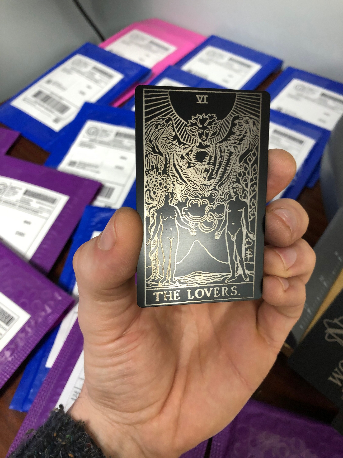 hand holding Metal The Lovers Tarot card laser engraved in matte black/silver in front of many mailer packages