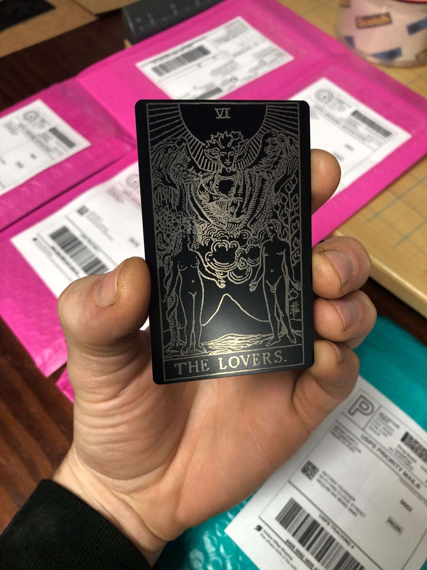Hand holding a metal tarot card with 'The Lovers' symbol against a pink box background