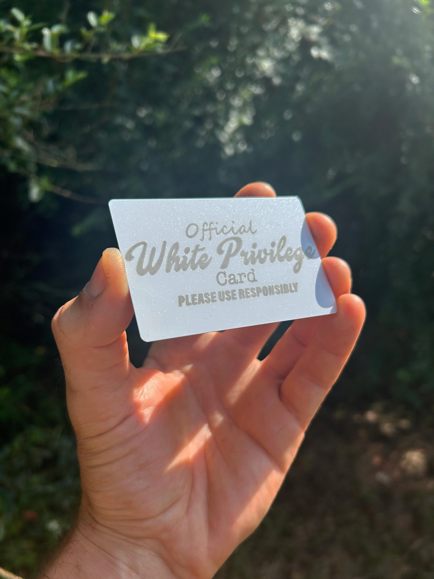 Hand holding a metal wallet insert card with 'Official White Privilege Card' sarcastic pop culture reference against a blurred natural background