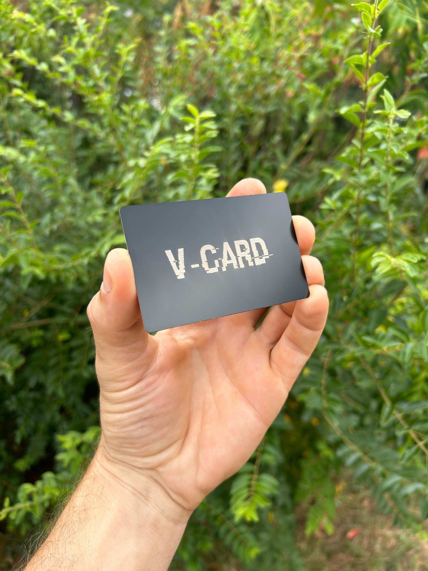 Hand holding a matte black metal card with 'V-CARD' text against a green background