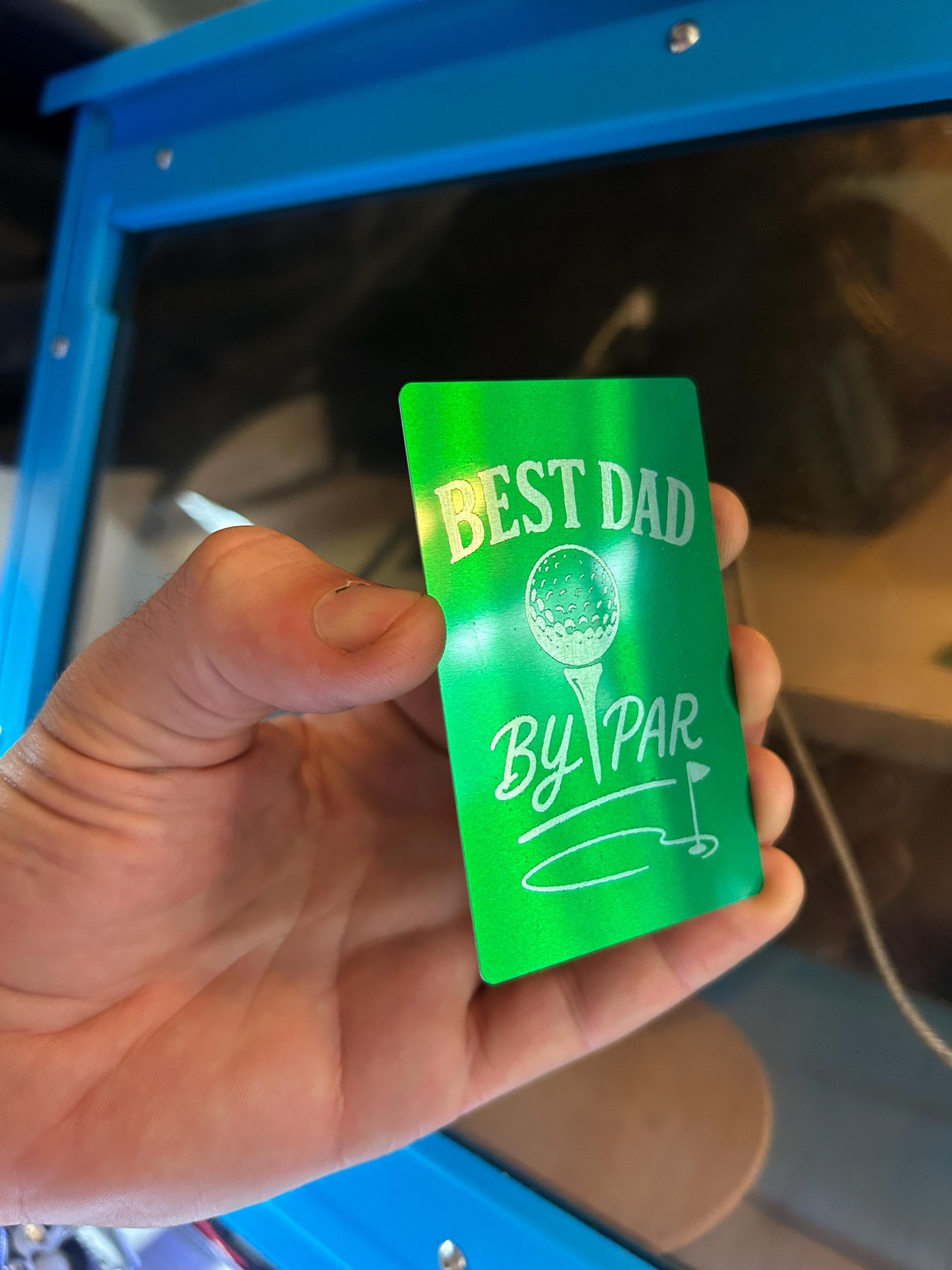 Hand holding a laser engraved metal green card with 'Best Dad By Par' text against a blue background