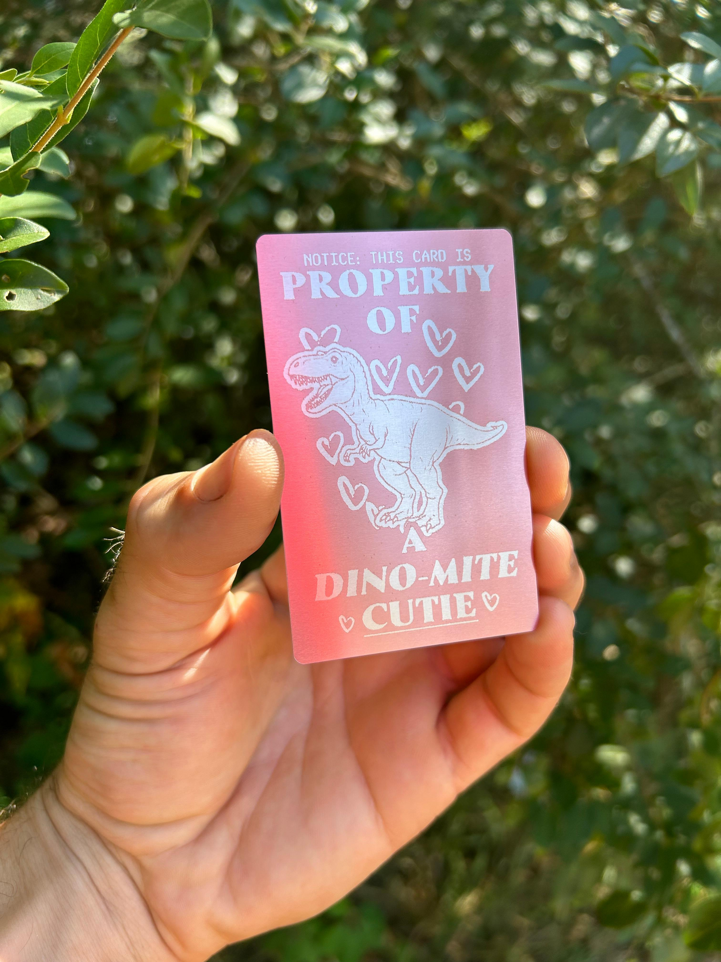 Hand holding a rose gold metalcard with a romantic dinosaur design and text against a green background