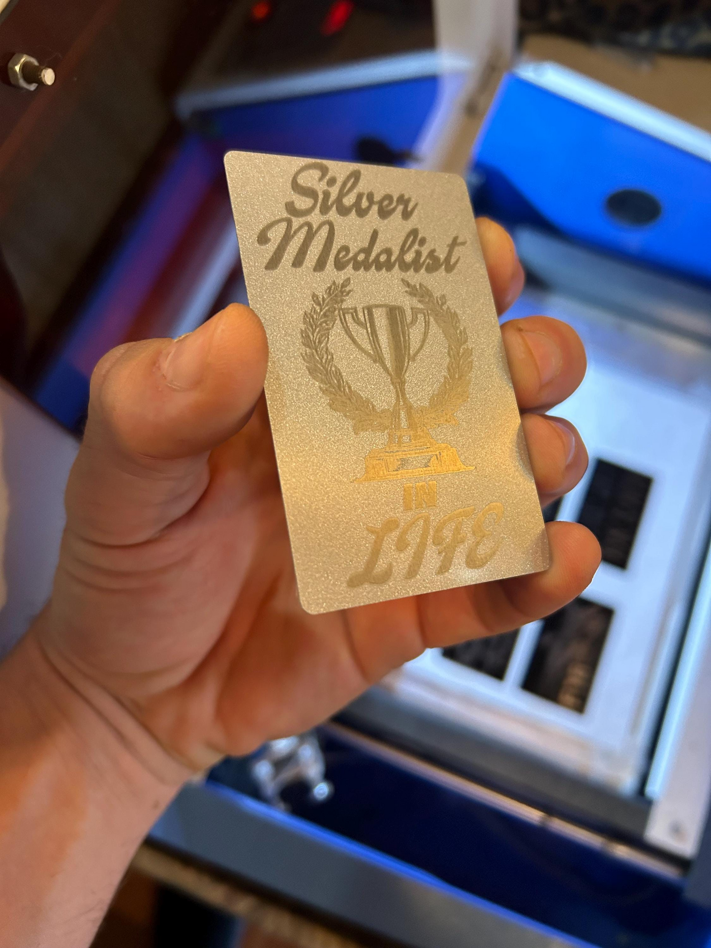 Hand holding a metal 'Silver Medalist In Life' sarcastic funny gift badge with a trophy design against a blurred background.