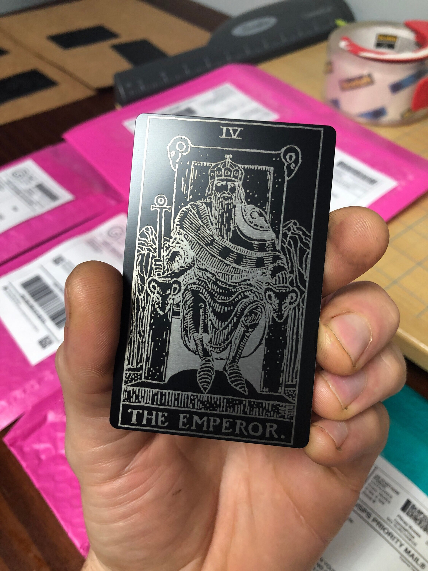 Hand holding a tarot card titled 'The Emperor' against several mailing packages on a blurred background.