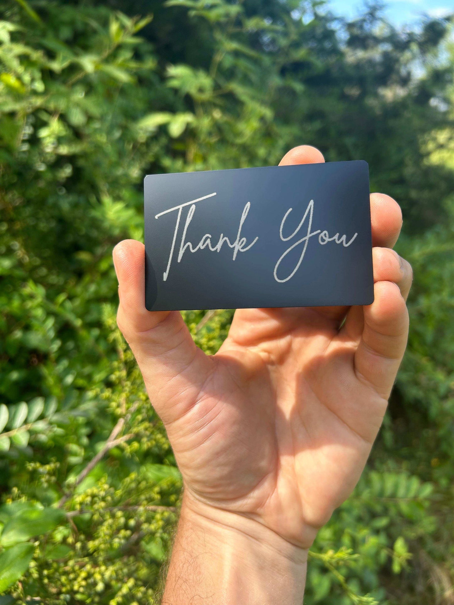 hand holding a metal laser engraved keepsake card insert that says thank you in laser-engraved silver text against a green nature background
