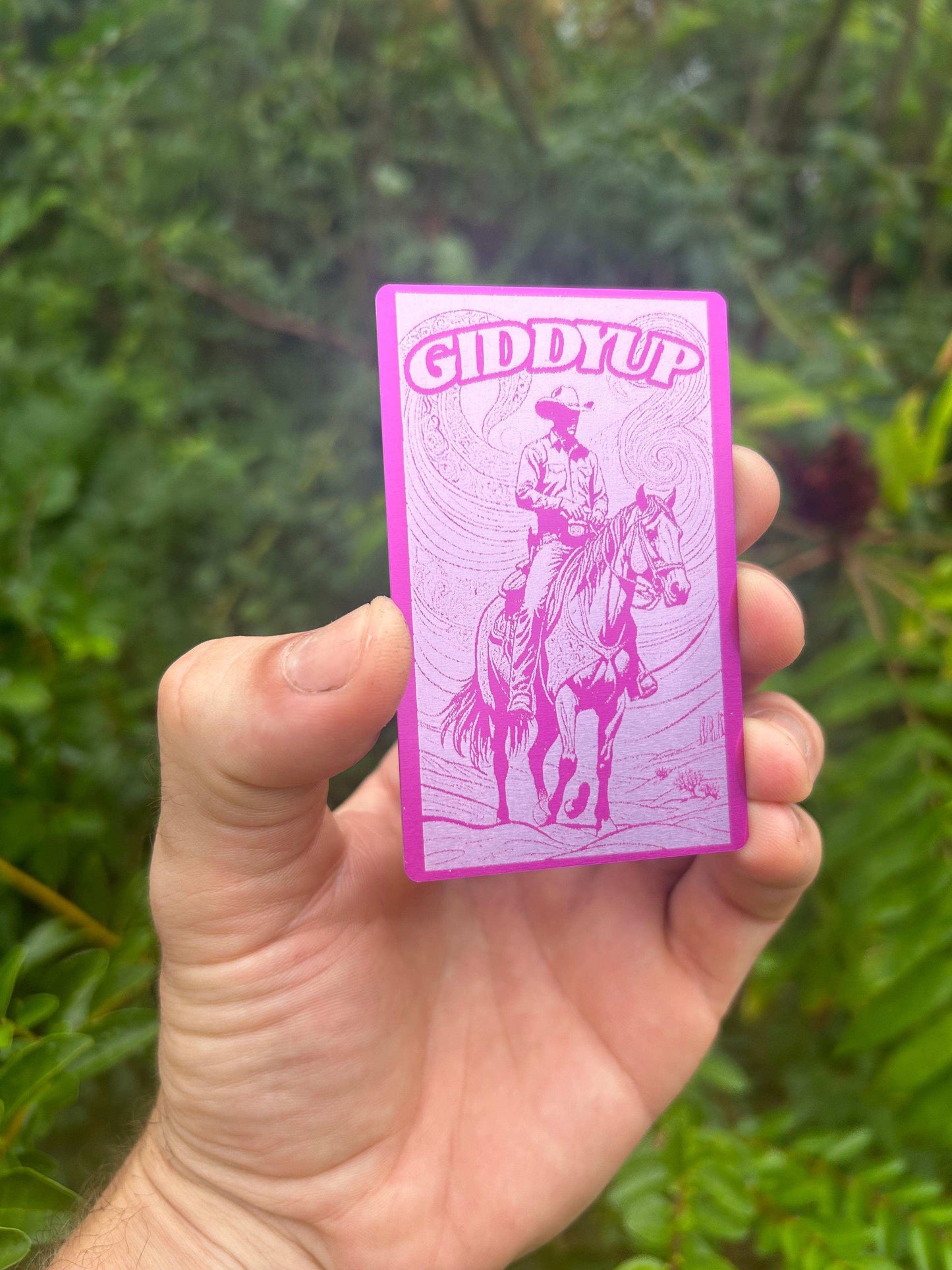 Hand holding a purple/silver card with 'Giddyup' and a rodeo rider illustration against a green background