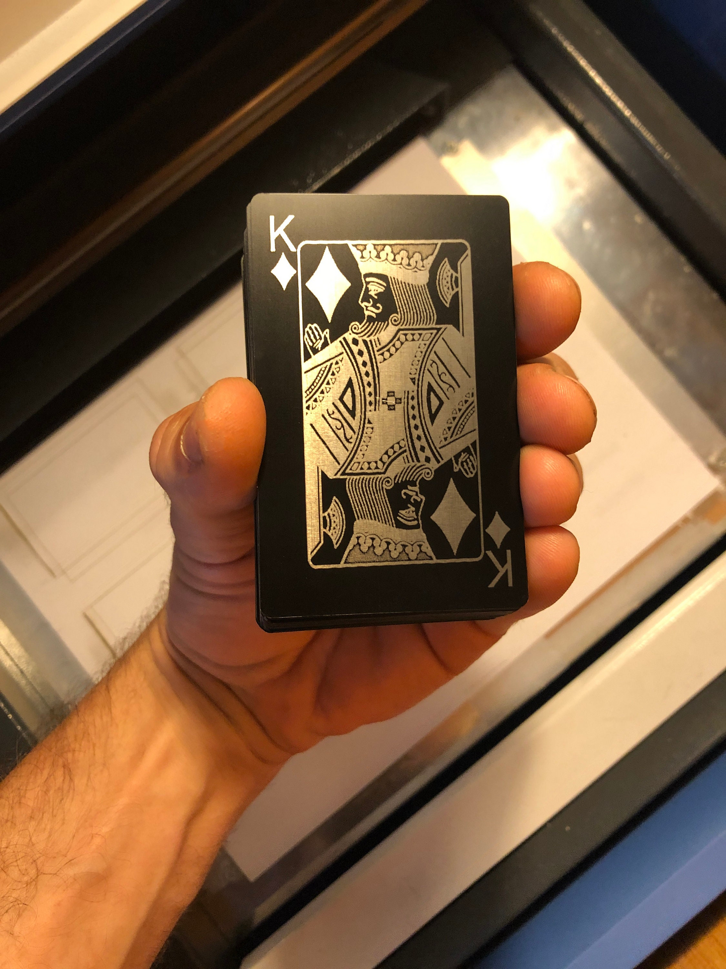 Hand holding deck of metal playing cards in black/silver with a laser engraved King of Hearts design on top, in front of a blurred machine background
