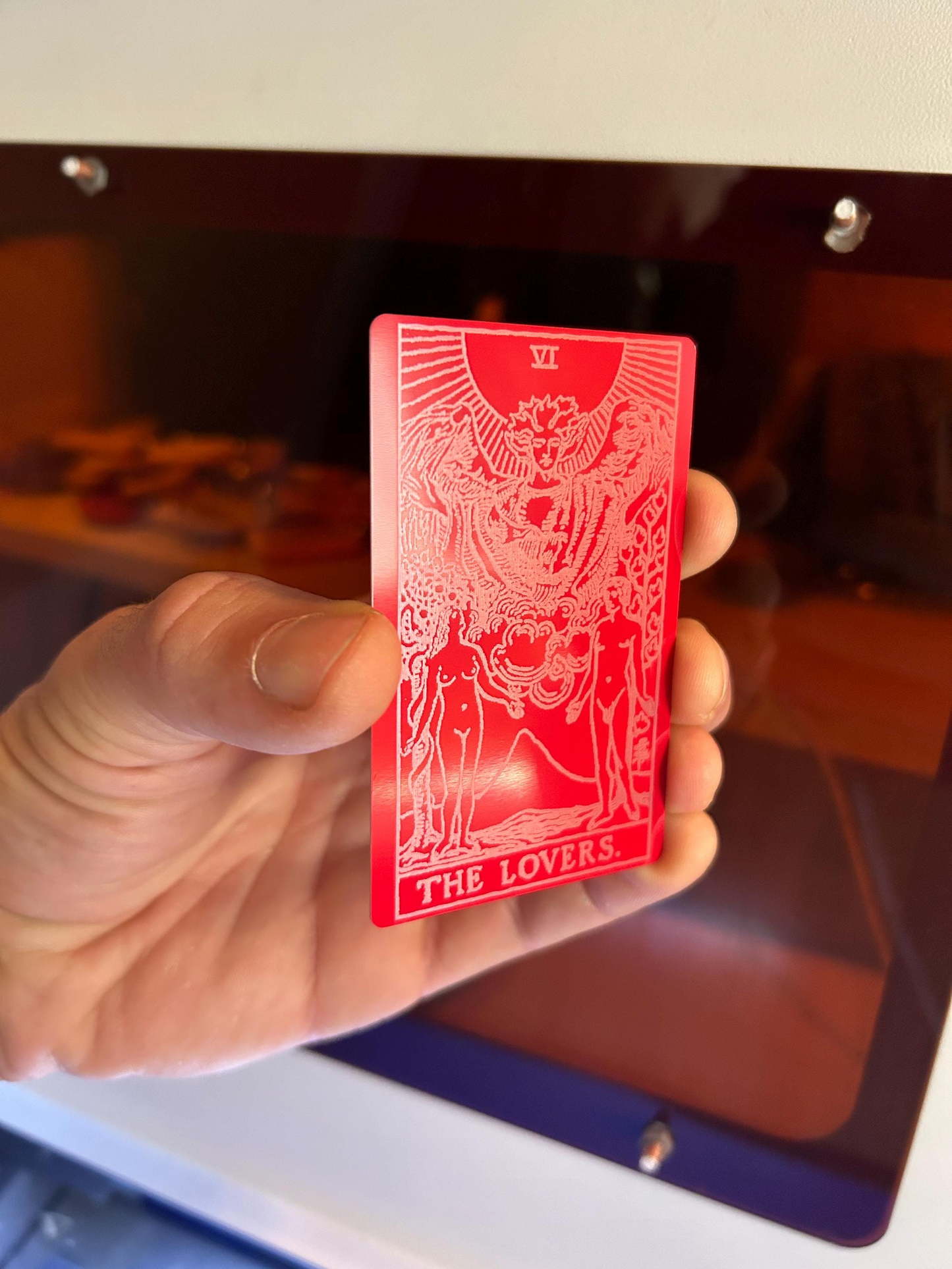 Red/silver metal tarot card titled 'The Lovers' held by a hand with a blurred background