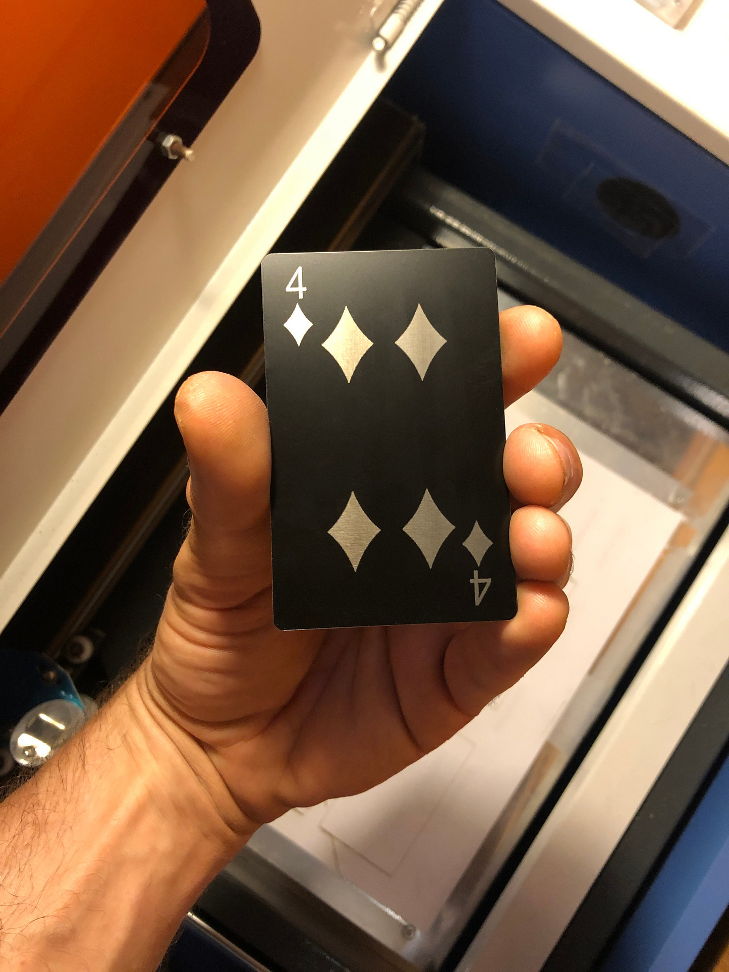 Hand holding a 4 of diamonds laser engraved playing card in front of a blurred machine