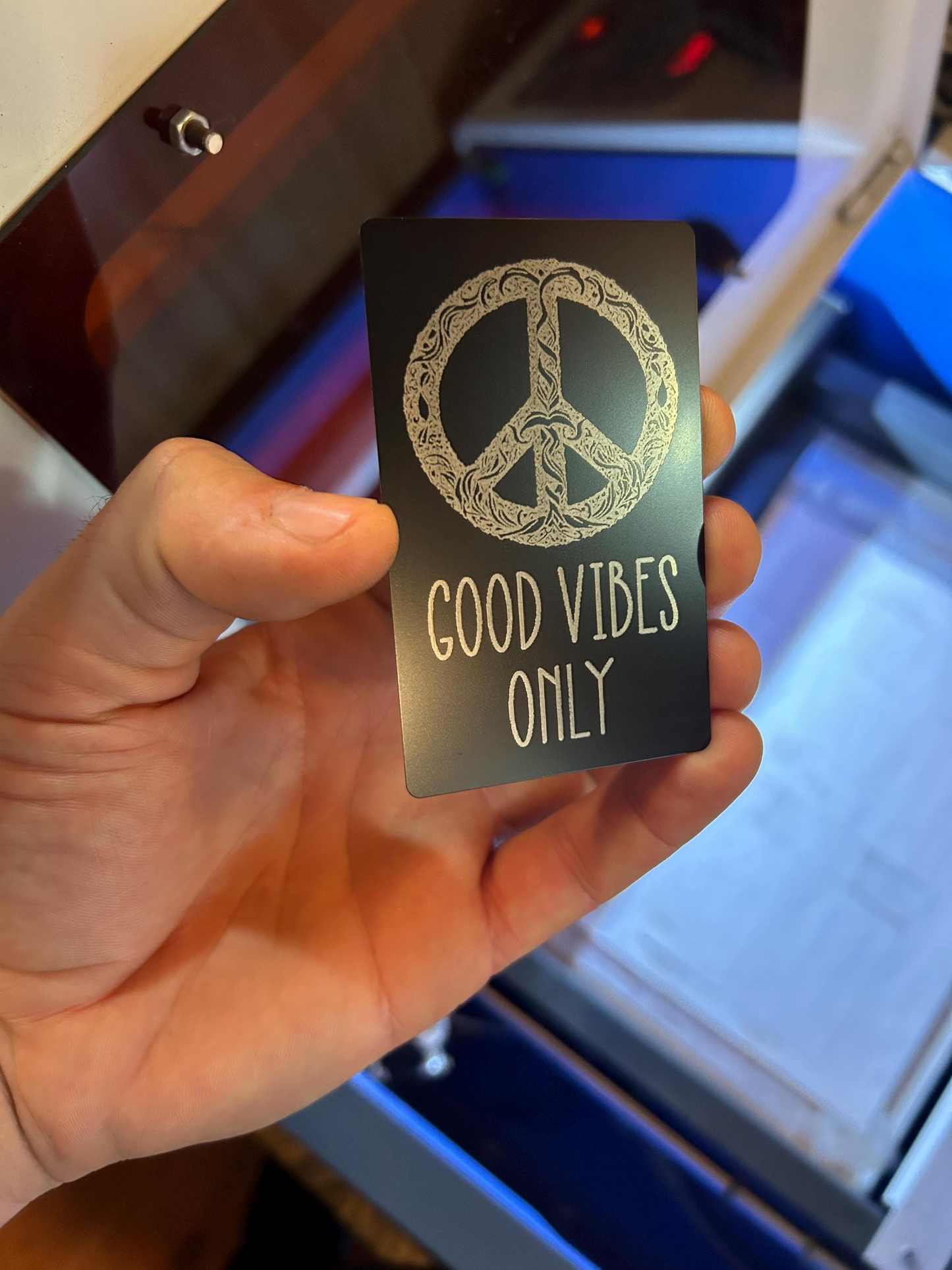 Hand holding a metal gift card with peace symbol and 'Good Vibes Only' text in front of a blurred background.