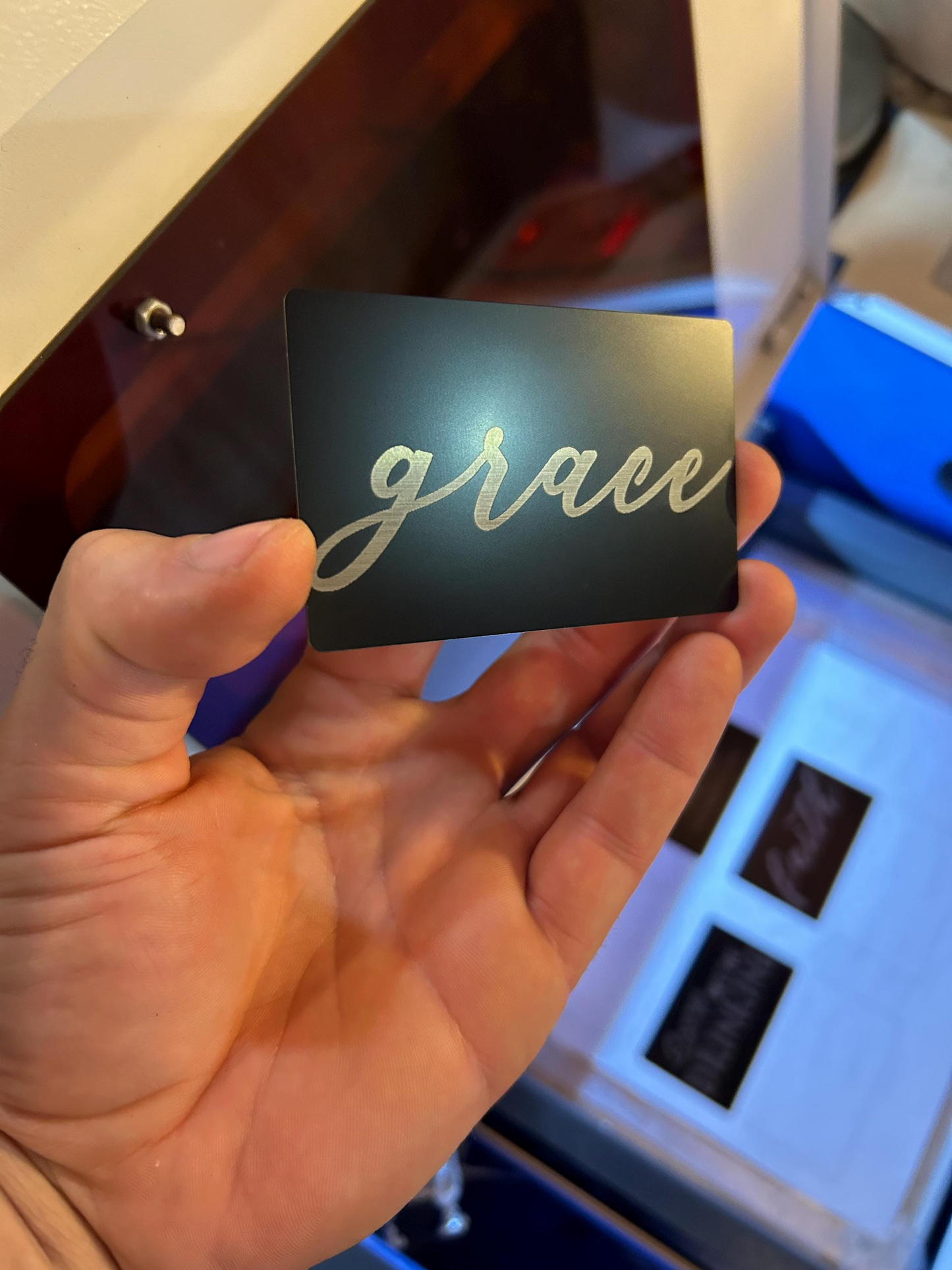 Hand holding a metal black/silver bookmark card with 'grace' laser engraved in cursive on a blurred background