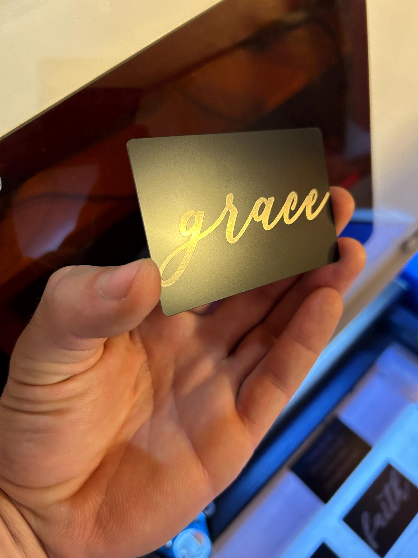 Hand holding a laser engraved black/silver metal keepsake card with 'grace' typography text against a blurred background