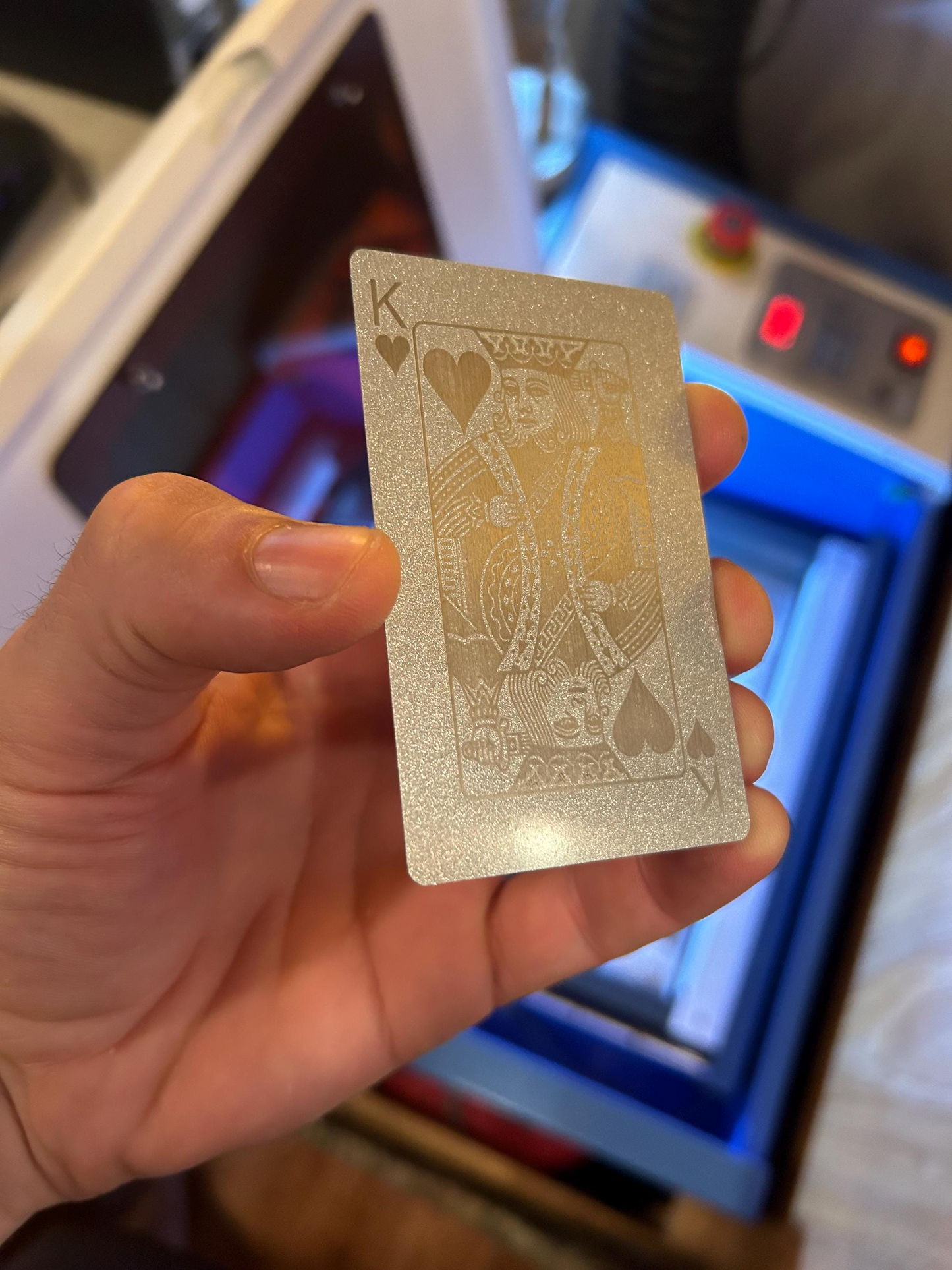 Hand holding a silver on silver playing card with a King of Hearts laser engraving in front of a machine in the background