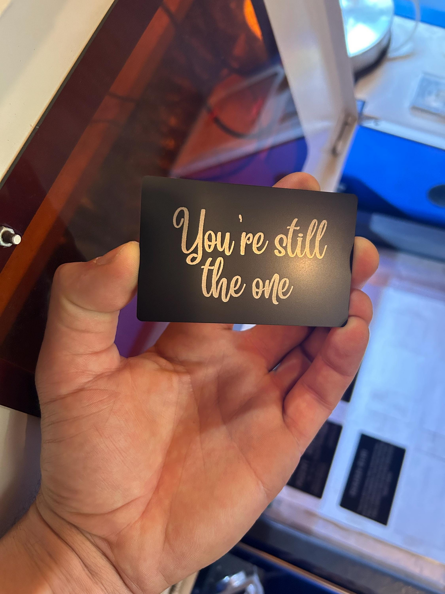 Hand holding a black metal card with 'You're still the one' in silver text against a blurred indoor background.