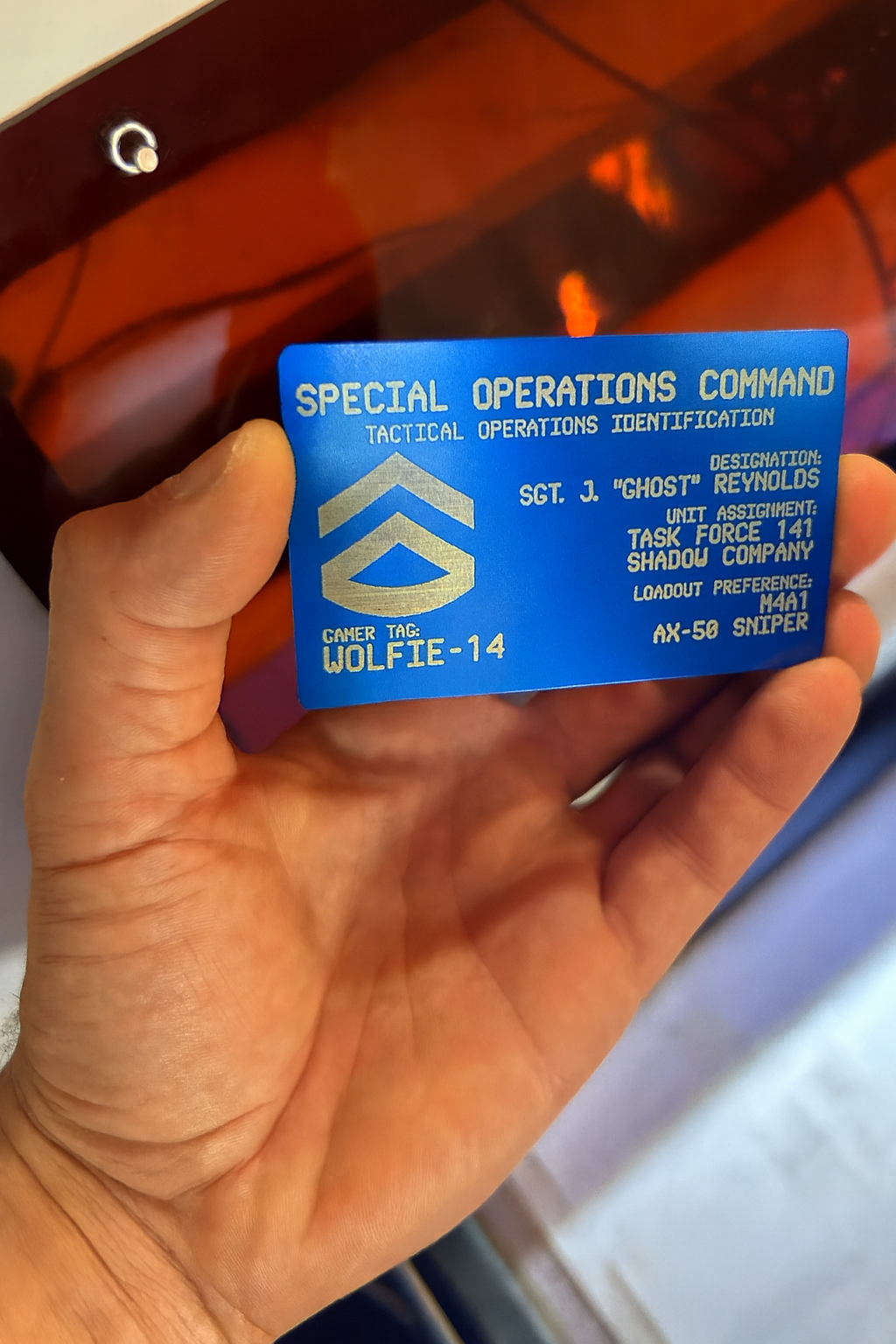 Hand holding a blue/silverFPS gamer identification card with personalized game text on a blurred machine background
