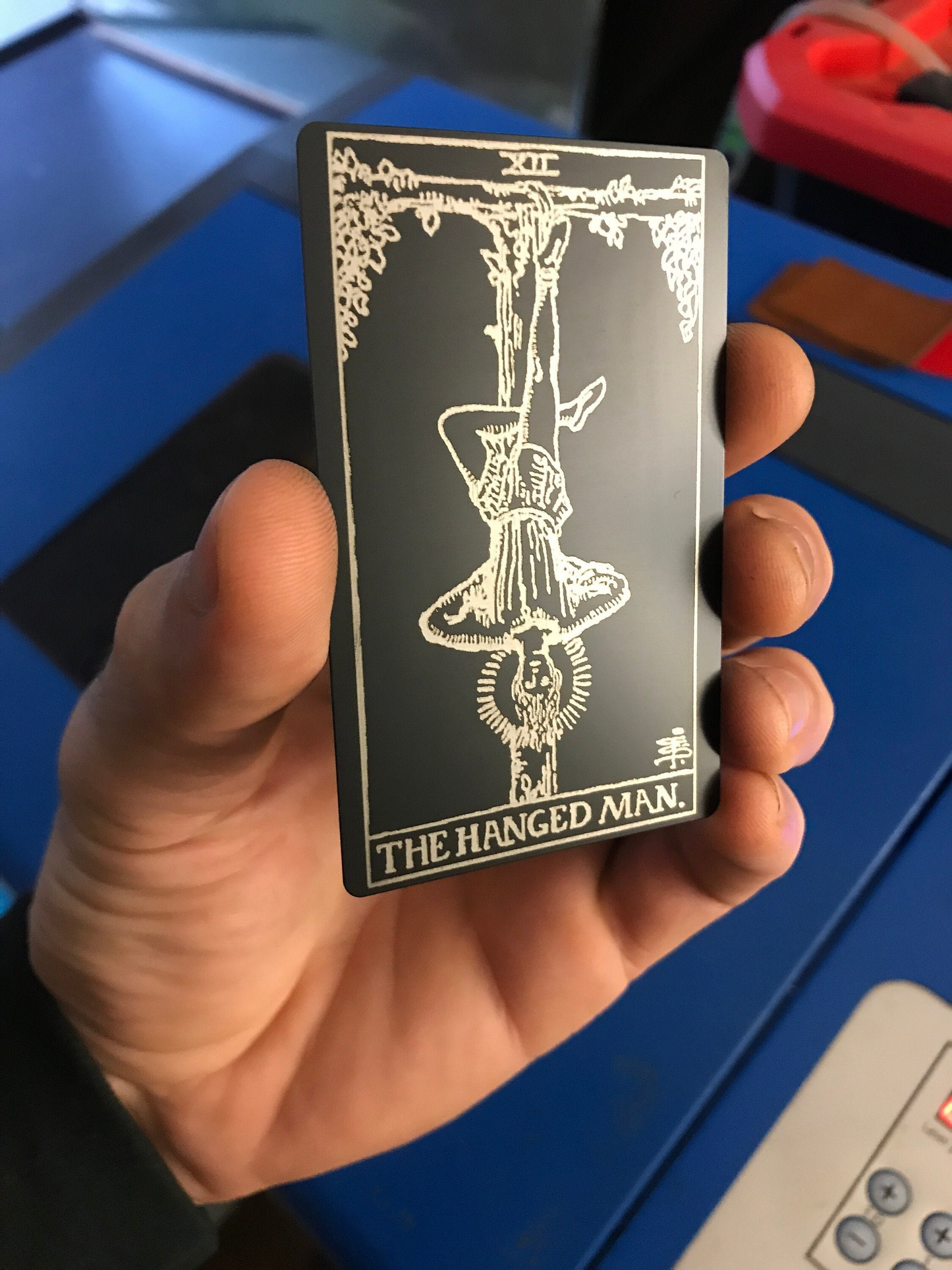Hand holding a metal laser engraved tarot card with 'The Hanged Man' design against a blue background.