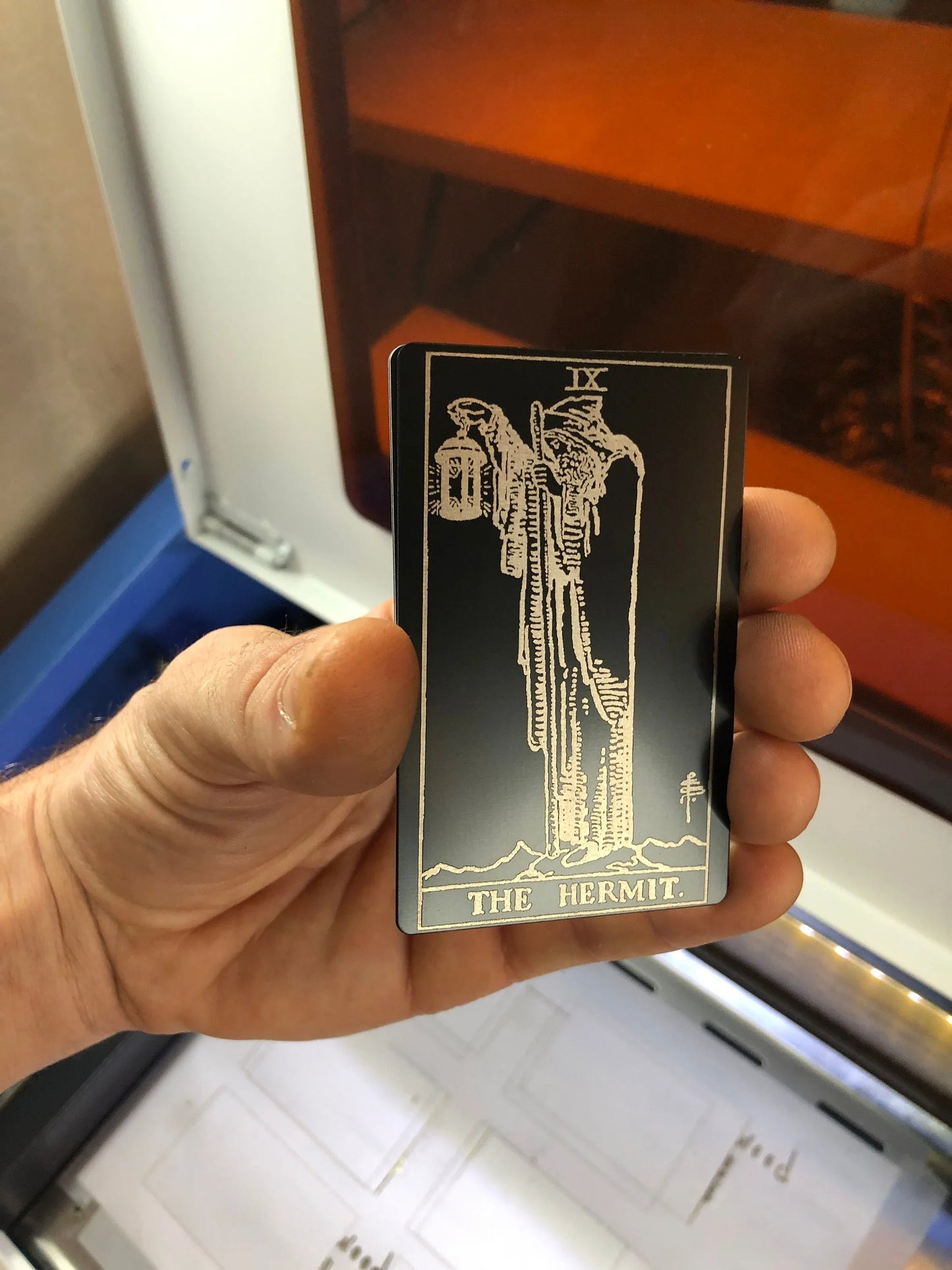 Hand holding a metal tarot card with 'The Hermit' illustration against a reflective surface.