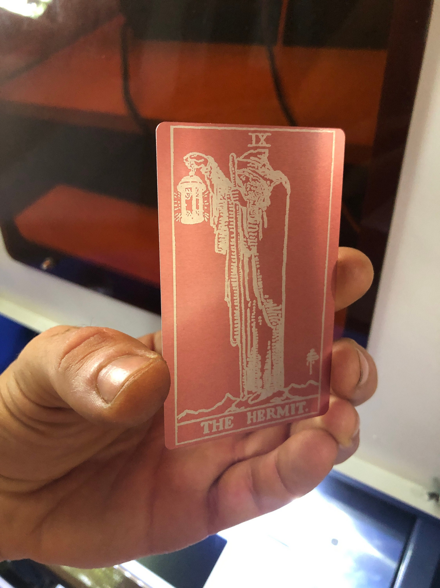 hand holding a rose gold and silver laser engraved metal tarot card, with The Hermit design -- made from anodized aluminum, held in front of a blurred laser cnc machine