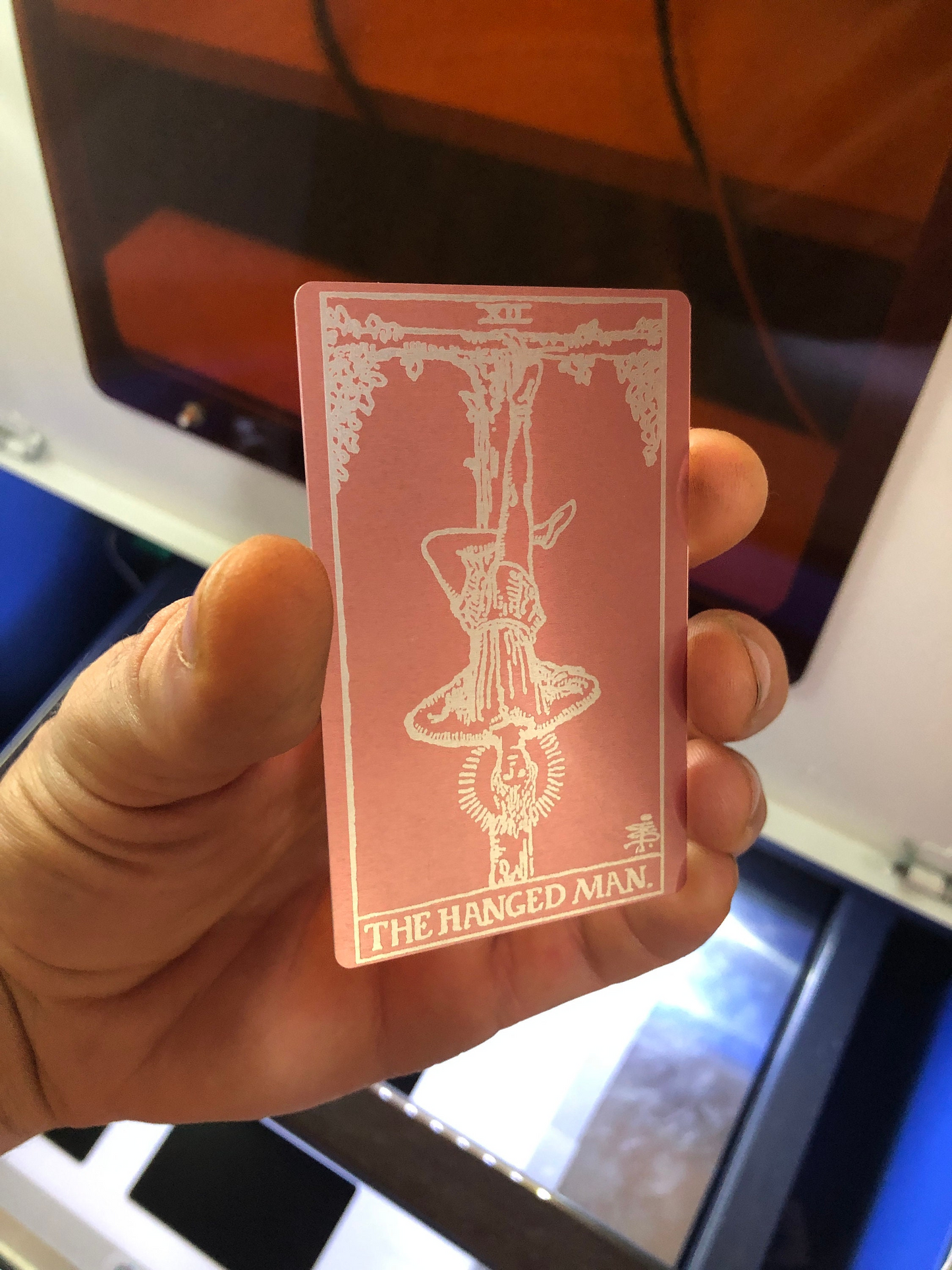 Hand holding a rose gold/silver metal tarot card with 'The Hanged Man' design against a blurred background