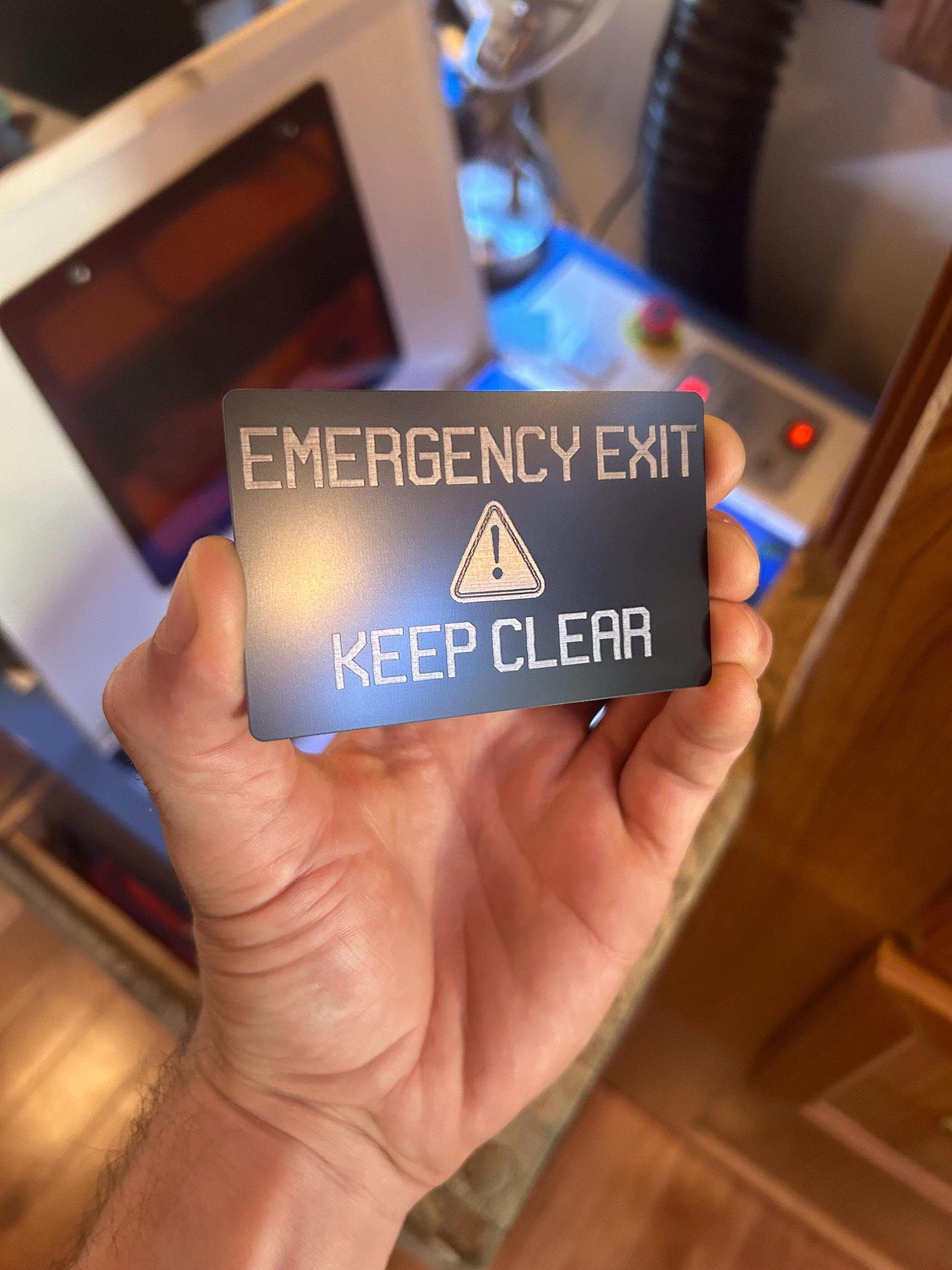 Hand holding a 'Emergency Exit Keep Clear' sign in front of a blurred industrial machine background.
