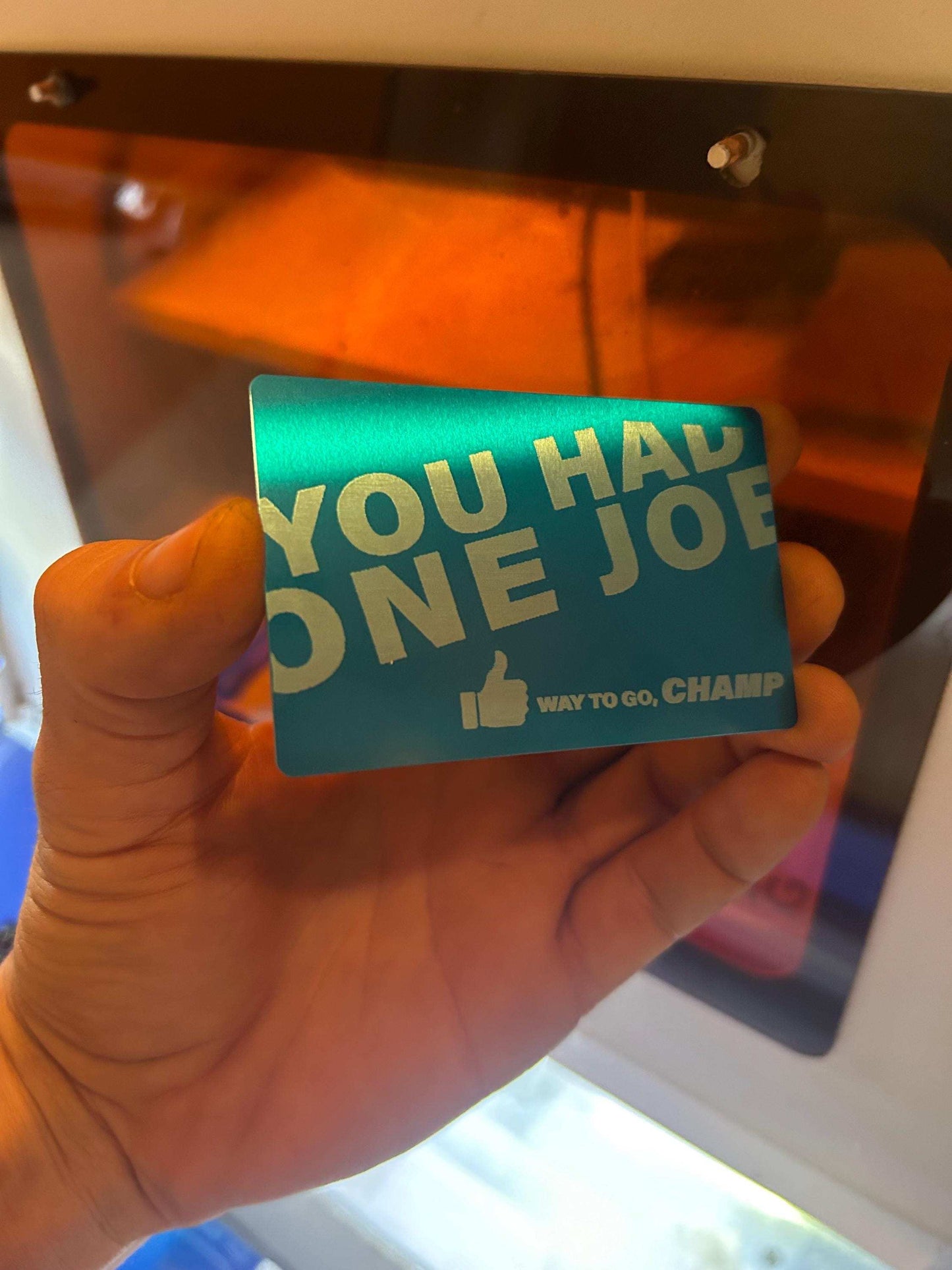 Hand holding a blue/silver engraved keepsake card with humorous text in front of a reflective surface