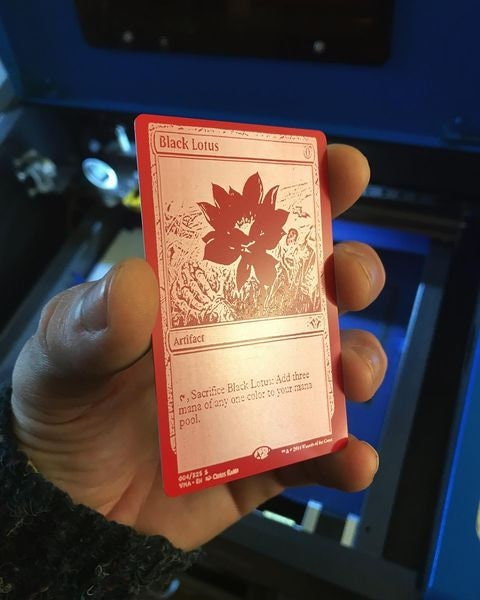 Hand holding a red and silver laser engraved gift card with a black lotus trading card design against a laser engraving machine background