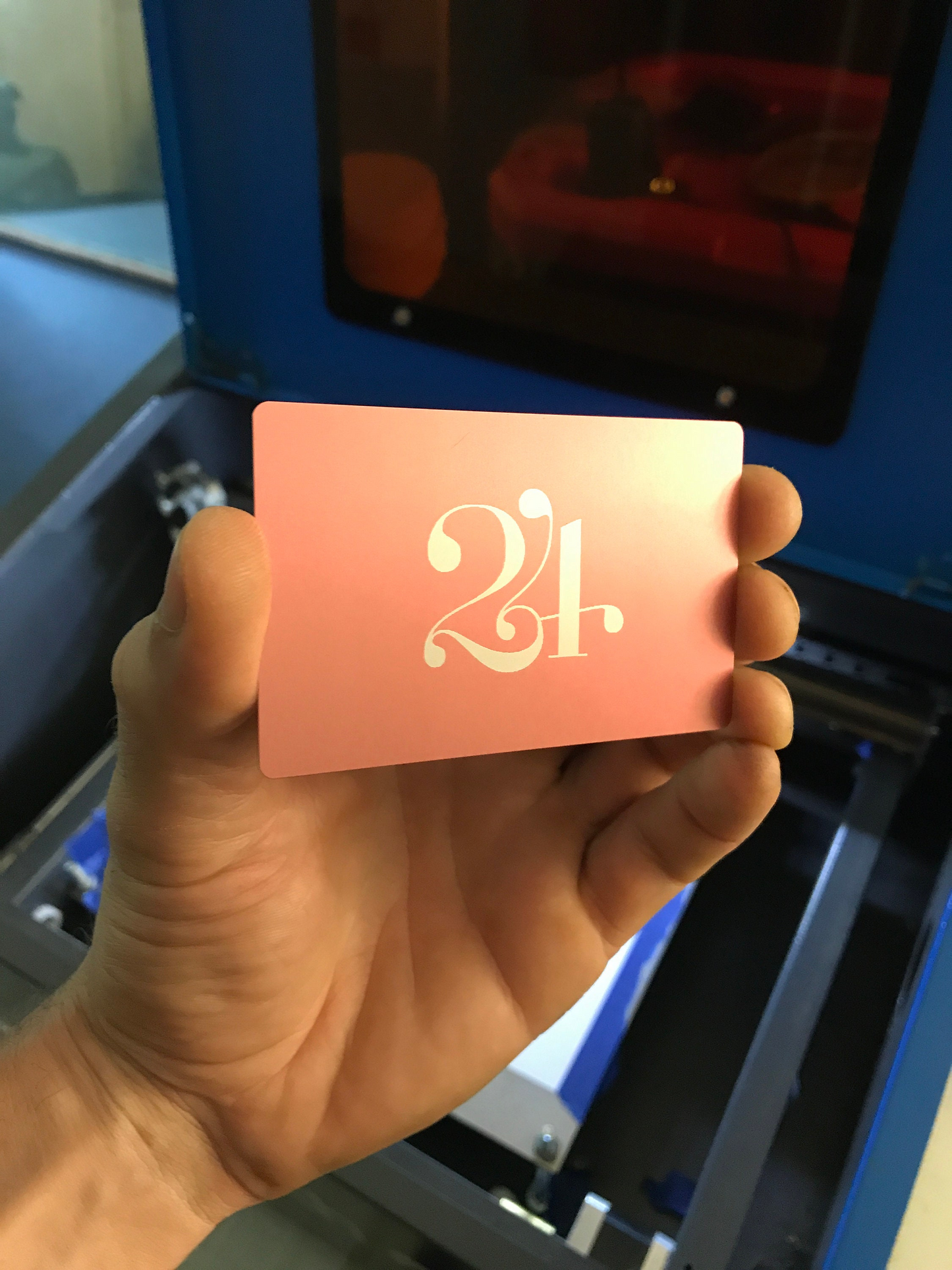 Hand holding a pink metal business card with personalized '24' on it in front of a cnc machine.