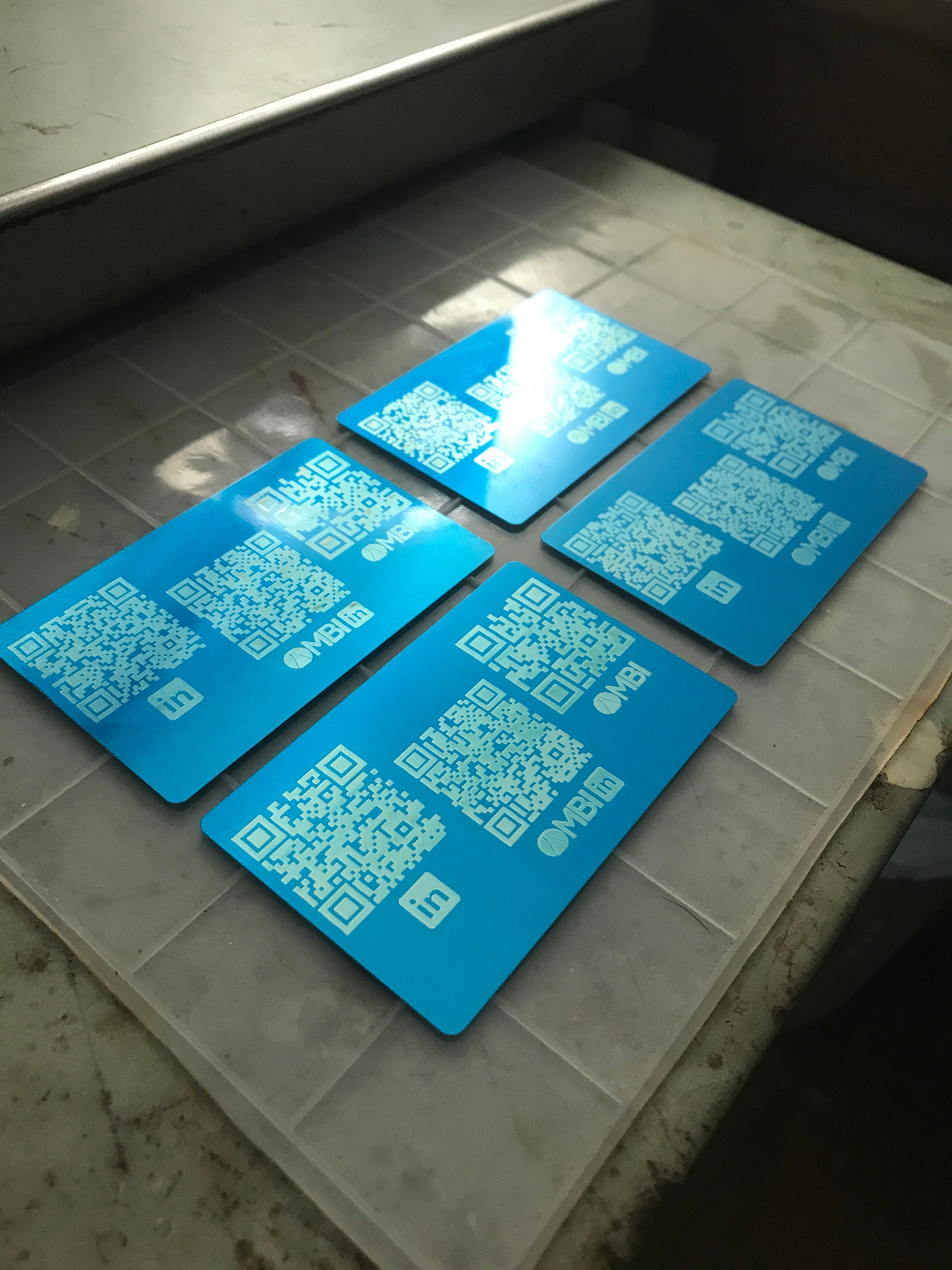 Four blue/silver laser engraved metal business cards with scannable QR codes for a small business promotion on a textured surface