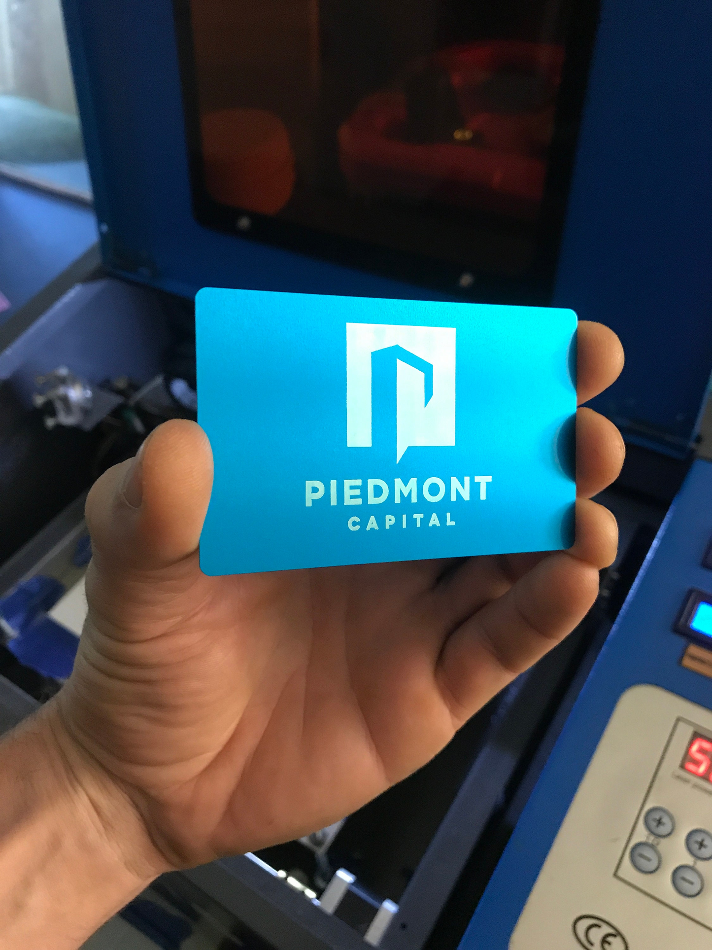 Hand holding a blue and silver metal business card with custom business branding engraved in front of a cnc machine.