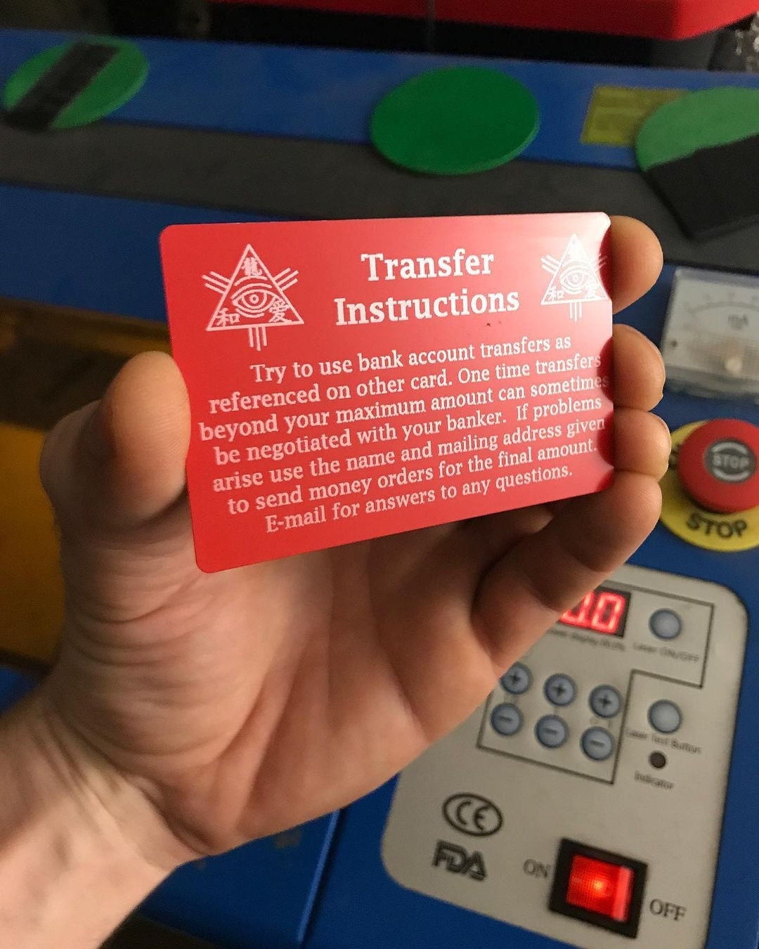 Hand holding a red/silver metal business card with laser engraved customized text of transfer instructions in front of a laser machine control panel.