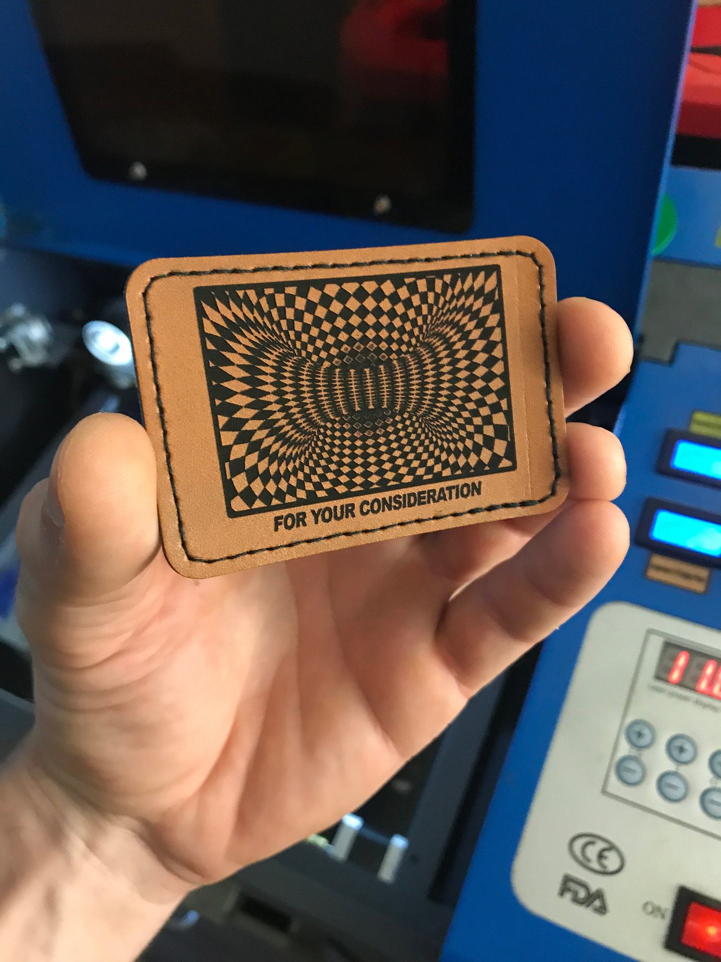 Hand holding a brown leather patch with black design and text in front of a blue machine.