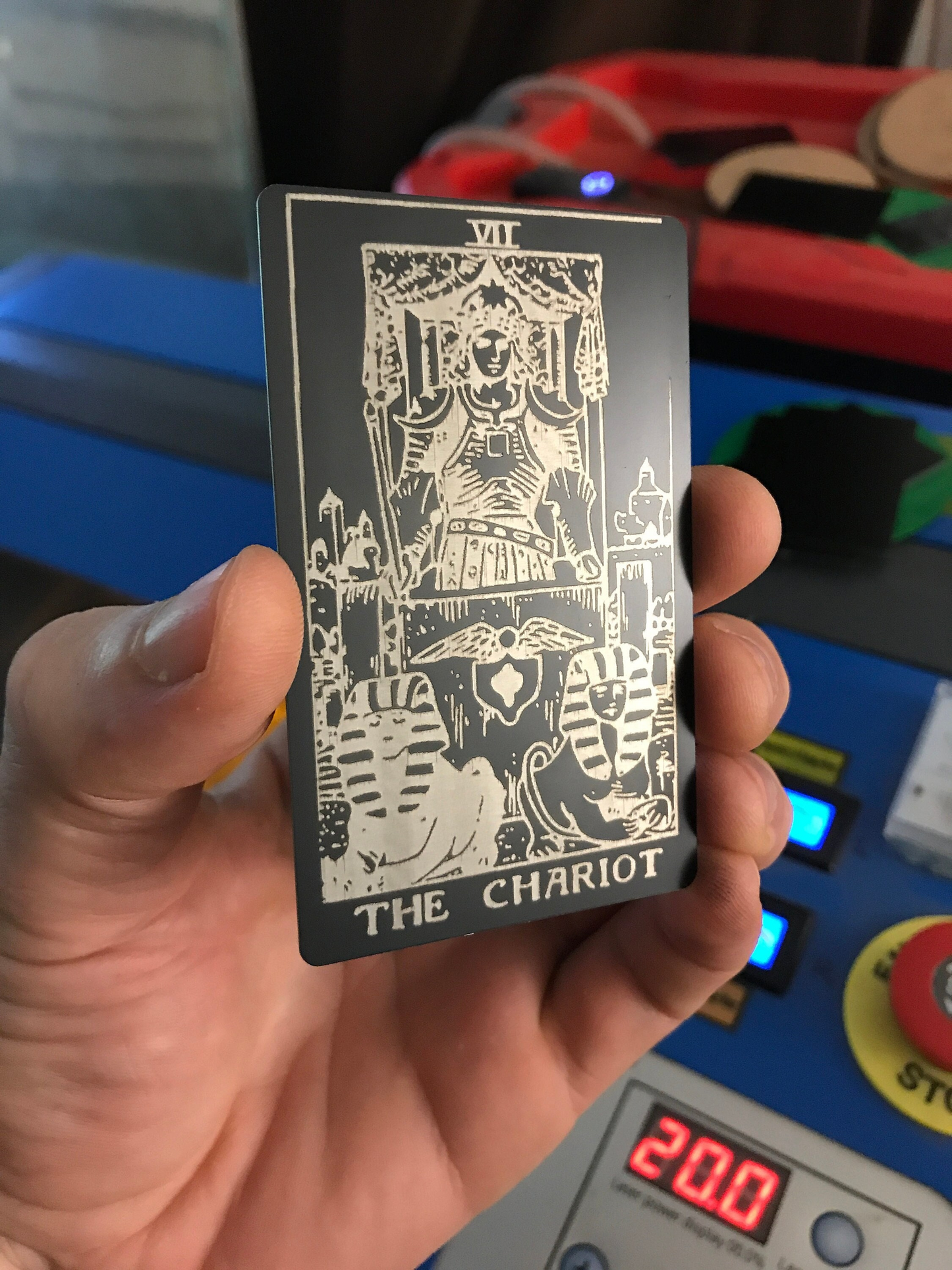 Hand holding a black/silver metal laser engraved tarot card titled 'The Chariot' in front of a colorful background.