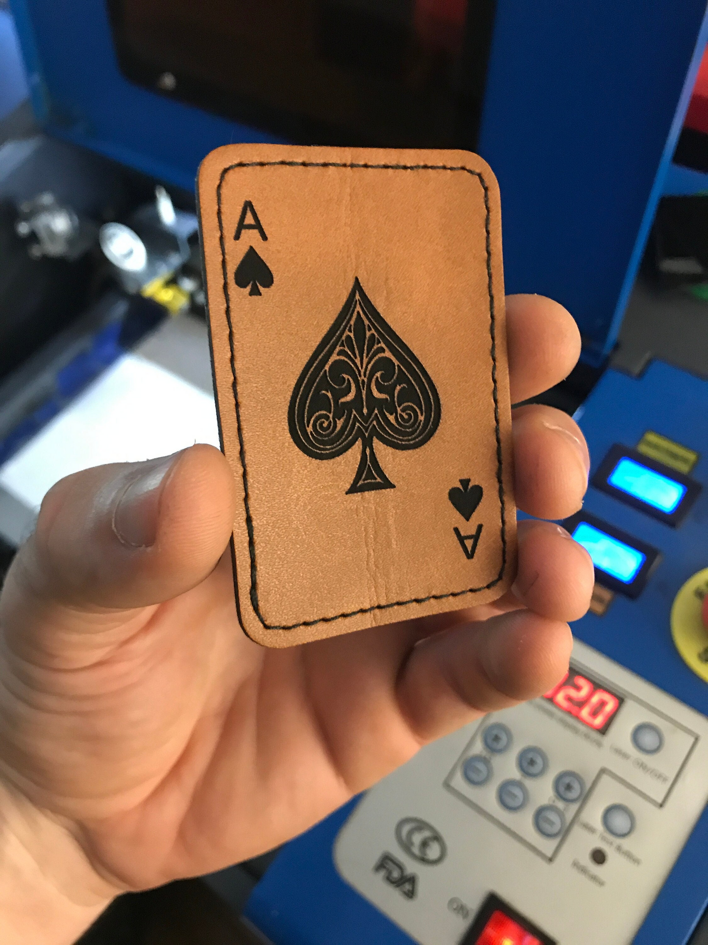 Hand holding a leather patch with an ace of spades design in front of a machine.